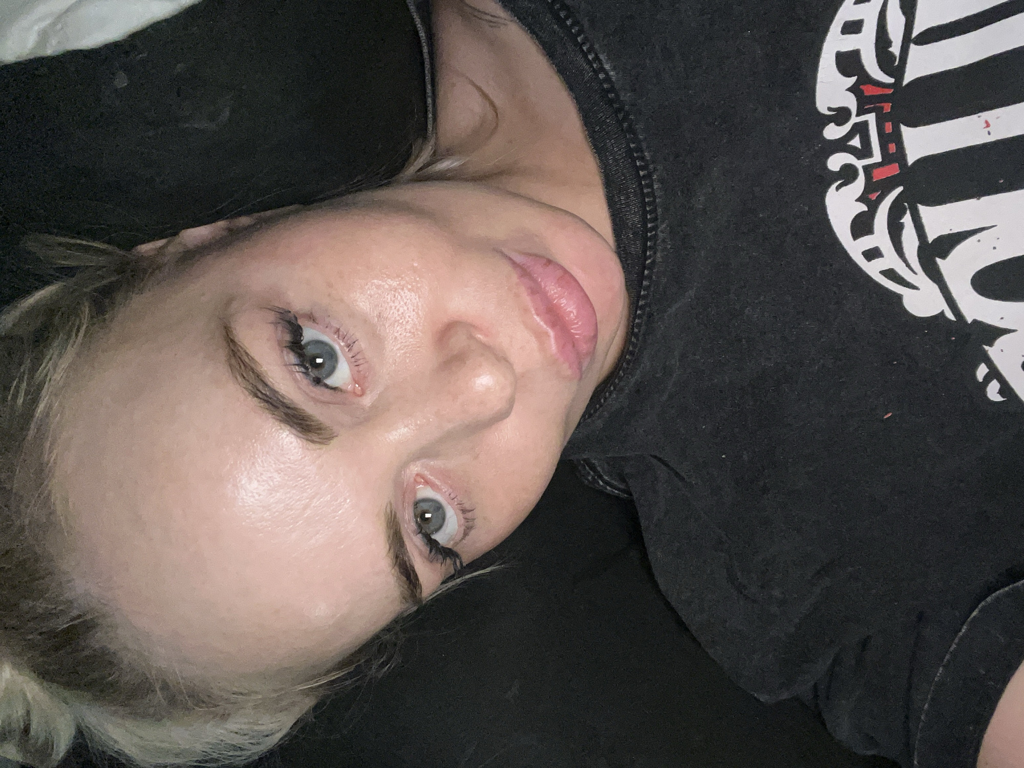 A close-up, low-angle shot shows a young, fair-skinned woman with blonde hair styled in two buns. She is wearing a dark, possibly black, t-shirt. Her eyes are blue, and she has false eyelashes. Her lips are slightly parted, and she appears to be making a pouty expression. The lighting is dim, casting shadows on her face and the dark background. The woman is lying down, with her head tilted back slightly. The image does not provide any information about her location in a city.