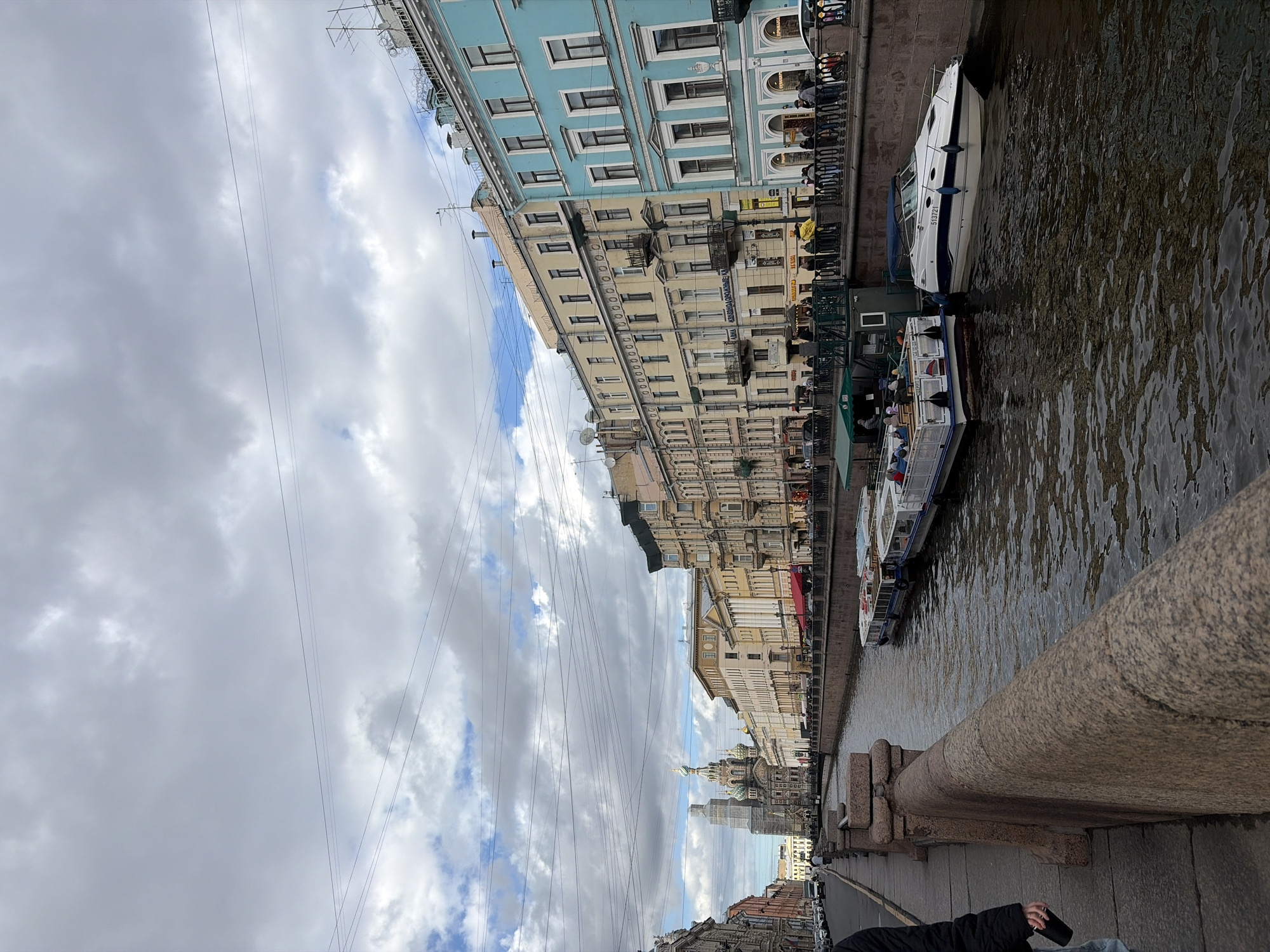 The image captures a daytime scene along a city canal in Saint Petersburg, Russia, viewed from a stone bridge. On the left side of the canal, a row of multi-story buildings lines the embankment. These structures display varied architectural styles and colors, predominantly light blue, yellow, and beige, with numerous windows and some balconies. Utility wires and antennas are visible against the sky above the buildings.
The canal itself, characterized by dark, rippled water, occupies the right side of the frame. Several boats are present on the water; a prominent white powerboat with blue accents is docked, displaying the number "513721." Further along, a smaller tourist boat carrying passengers can be seen. Small figures of people are visible both on the boats and walking along the canal's edge.
In the foreground on the right, a textured, speckled granite railing of the bridge from which the photograph was taken is clearly visible. The sky is largely overcast with a mix of white and grey clouds, though patches of blue sky are discernible, indicating a partly cloudy day. In the distance, the distinctive, colorful domes and spires of the Church of the Savior on Spilled Blood are visible, serving as a key landmark that identifies the location as Saint Petersburg. The scene depicts a typical busy city waterway.