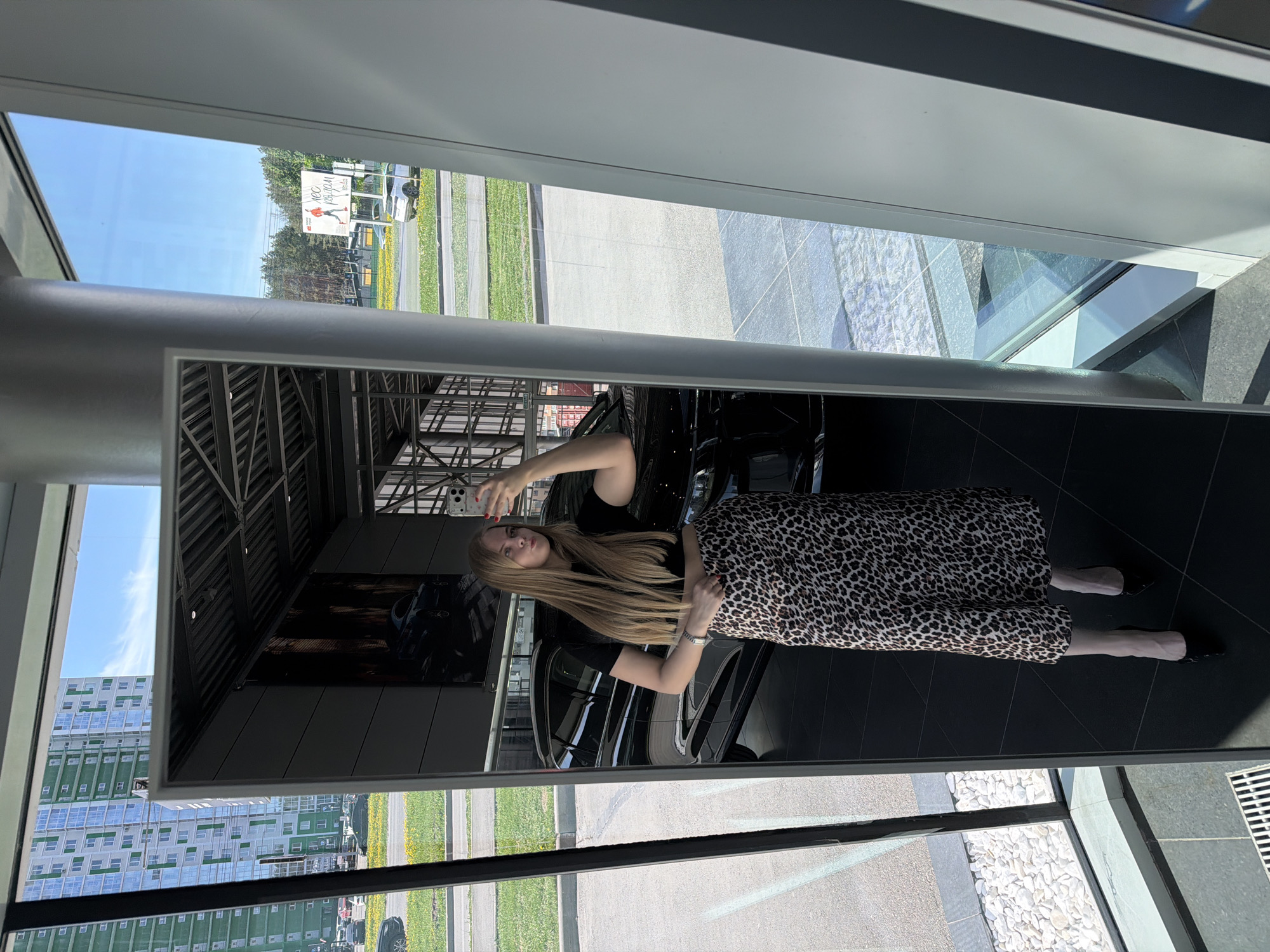 A woman stands in a showroom, reflected in a window. She is wearing a black top and a leopard-print skirt, with long blonde hair. Outside the window, there is a building that identifies the location as Izhevsk, Russia. Inside the showroom, several black cars are visible. The woman is holding up one hand, as if posing for the reflection.