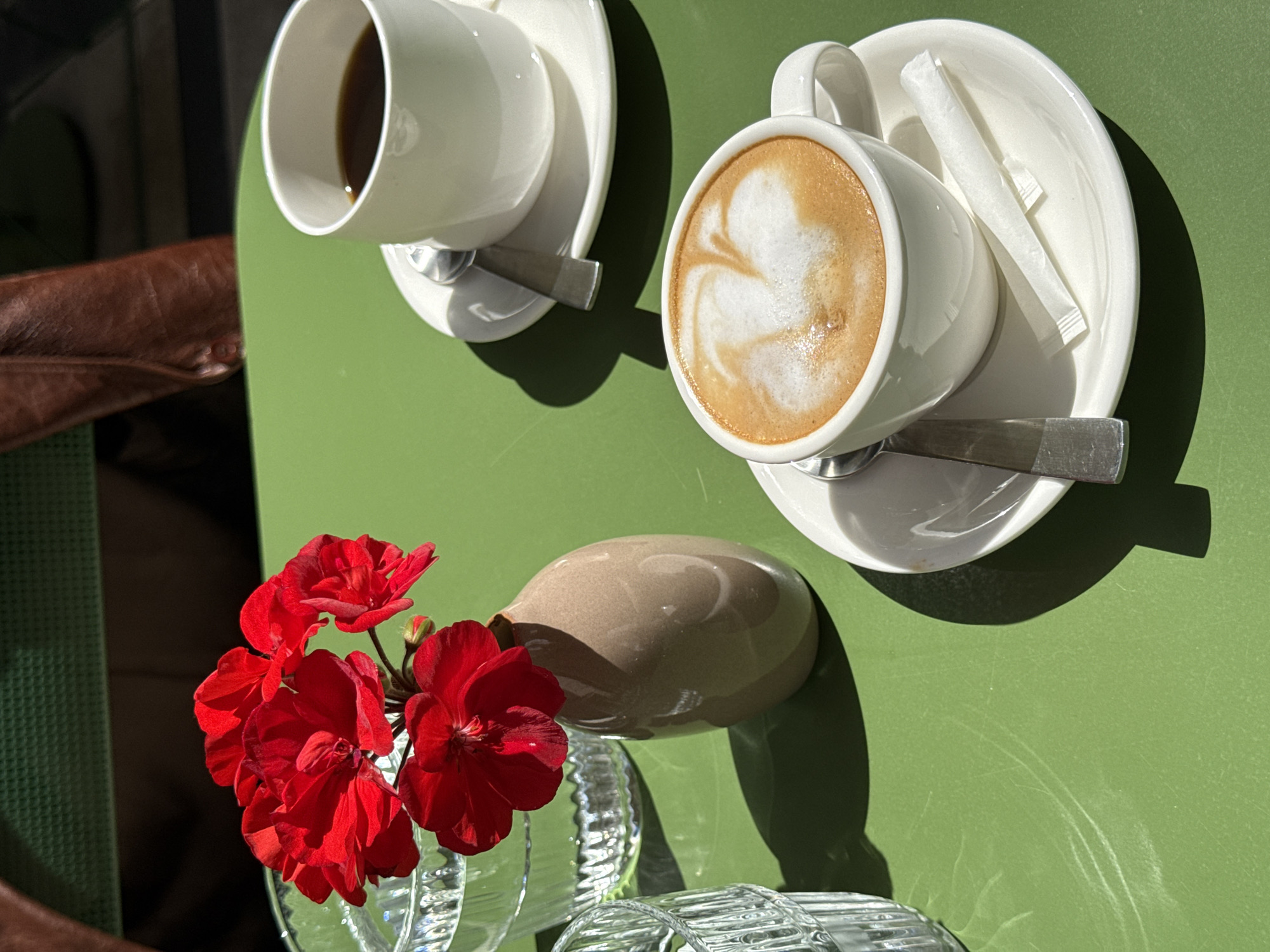 Two cups of coffee, one with black coffee and the other with a latte, are placed on a green table. Both cups sit on white saucers, each with a spoon and sugar packets next to the latte. A small vase of bright red geraniums is positioned nearby. The scene is set outdoors, likely on a patio or sidewalk café, on a sunny day, casting sharp shadows on the table. The location is Belgrade, Serbia.