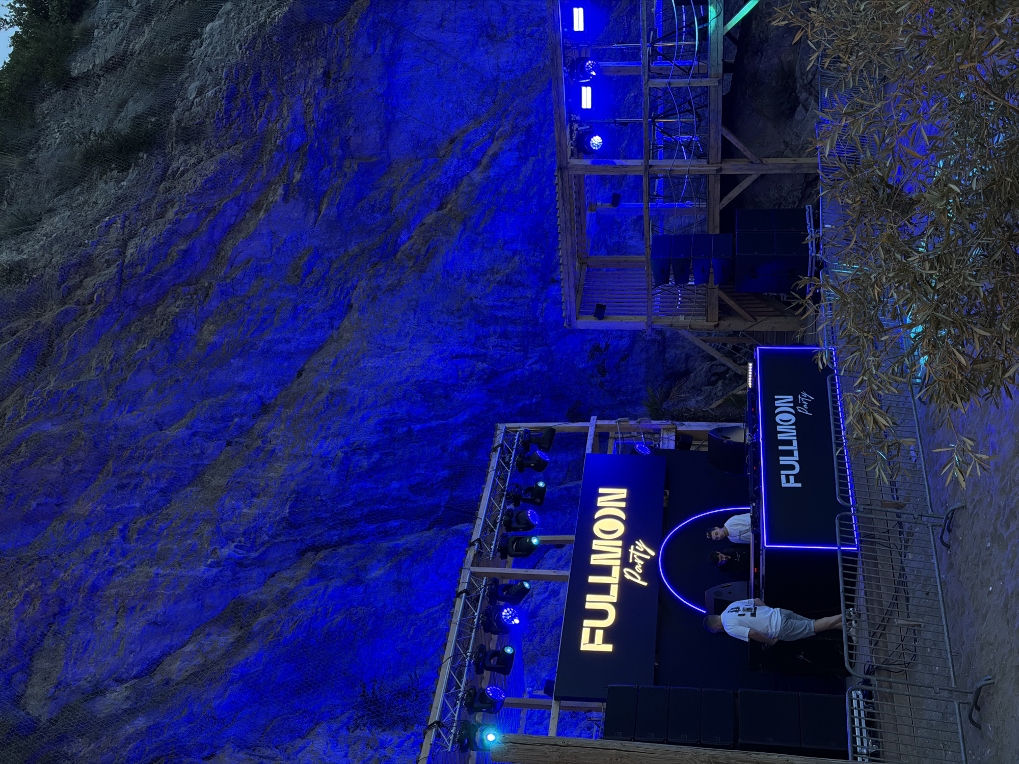 Captured in the city of Glavatičići, Montenegro, the image showcases a "FULLMOON Party" event set against a rugged cliff face, illuminated with bright blue lighting. Two wooden structures are visible, one appearing as a stage setup for the party, and the other a multi-level platform. Speakers, light fixtures, and signage bearing the event name are mounted on the stage structure. Behind the stage, two DJs can be seen performing. The lower portion of the stage is lined with a metal barrier in front of it, with what looks like tree branches on the side. The multi-level structure has blue lights positioned on each level. The cliff face itself is a prominent feature, with a protective mesh net over some parts of it. The environment is dark overall, accentuated by the bright blue lighting on the cliff and event structures. A person, wearing shorts and a t-shirt with number 44 on it, can be seen at the bottom of the image.