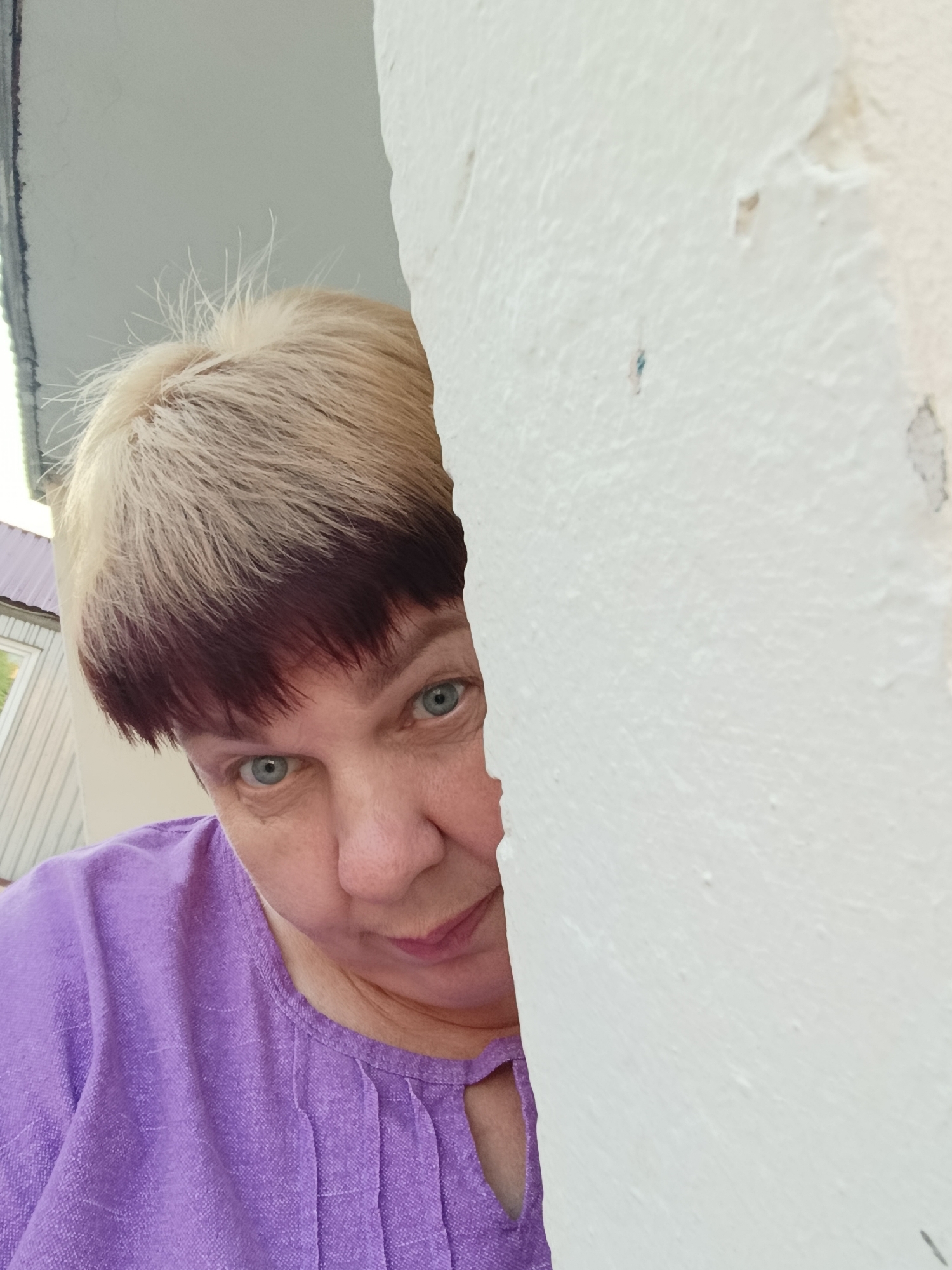 A woman with short, blond and dark-dyed hair peeks out from behind a white wall. She is wearing a purple top. The background shows a building with a light-colored facade and a metal roof. The image appears to be taken outdoors, possibly in a residential area of Svietlahorsk, Belarus.