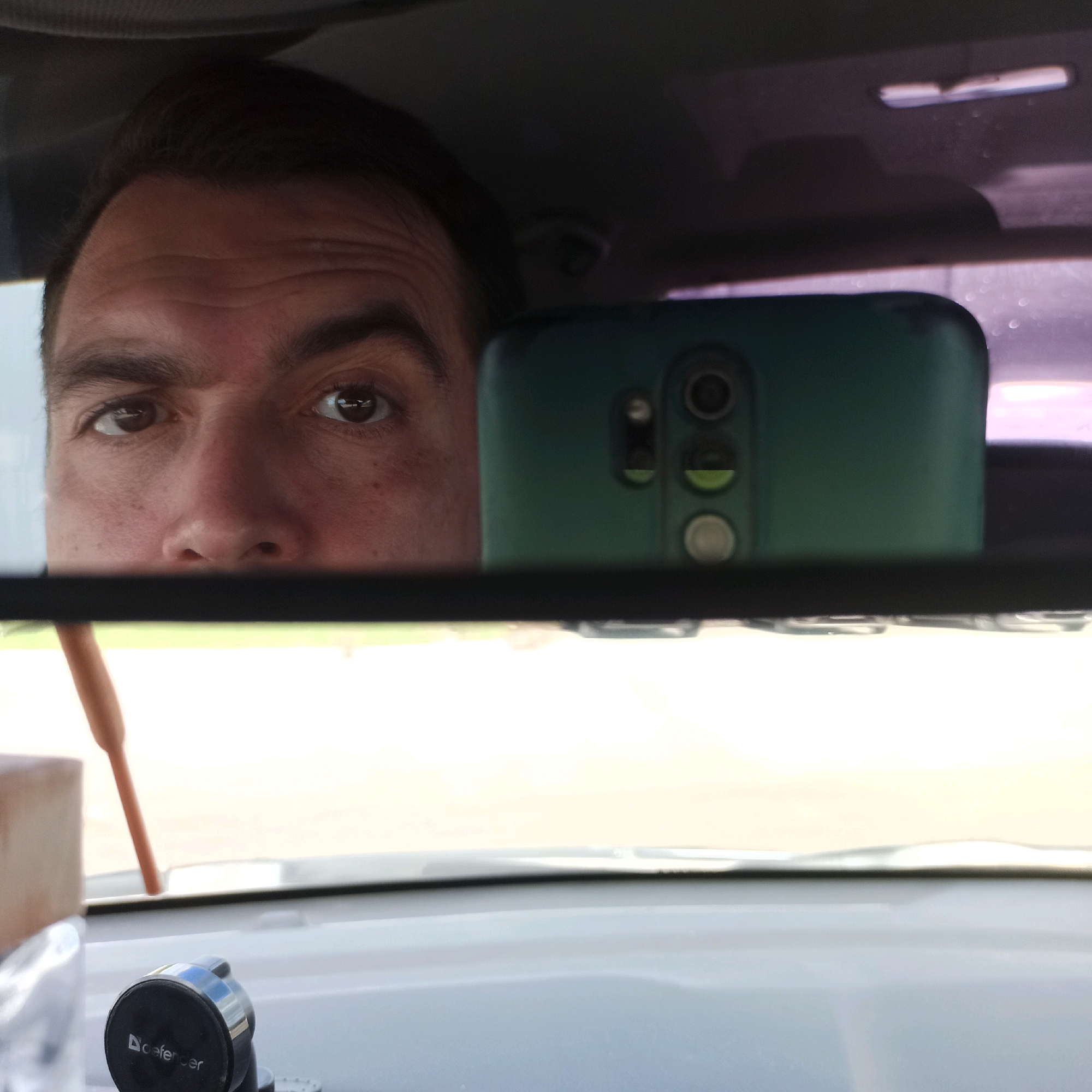 The image is a reflection in a car's rearview mirror, showing a man's face. He appears to be in the driver's seat, with his eyes looking directly at the camera. The background of the reflection shows a green smartphone. The car's interior is visible in the foreground, including a car mount with the brand "Defender" and a portion of the dashboard. The image quality is moderate, with some graininess and a slight blur. The overall impression is that of a candid selfie taken in a car. There are no indications of the location being Nizhny Novgorod, Russia; this information is not present in the visual data.