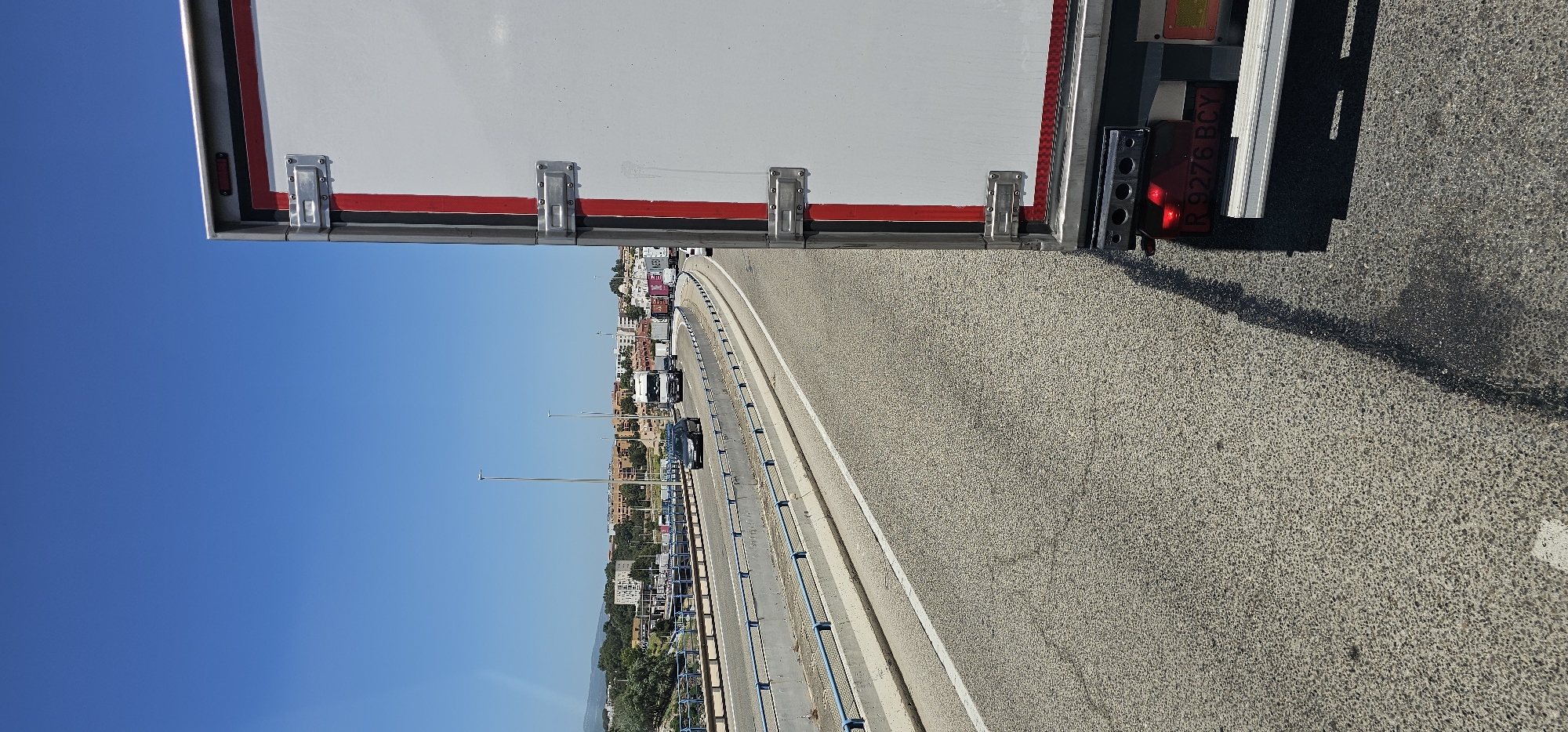 The image captures a sunny daytime scene from the perspective of a vehicle, looking along a curved road with traffic. The foreground is dominated by the side of a large white and red vehicle, likely a truck or trailer, extending across the top and right side of the frame. Attached to its rear, a red brake light cluster and a partially visible license plate are seen.
The road is a multi-lane asphalt surface, curving towards the right, with a clear white line delineating a lane. A blue metal guardrail runs along the side of the road. In the middle ground, several cars and trucks are visible in traffic, suggesting slow-moving conditions. Streetlights with tall poles are spaced along the roadside.
The background reveals a town or city built on hills, with numerous buildings under a clear, vibrant blue sky. Some green vegetation is also visible between the road and the distant buildings. The weather is bright and sunny.
Visible text on the license plate of the vehicle in the foreground reads "R 9276 BCY". The scene depicts a typical road environment in Algeciras, Spain.