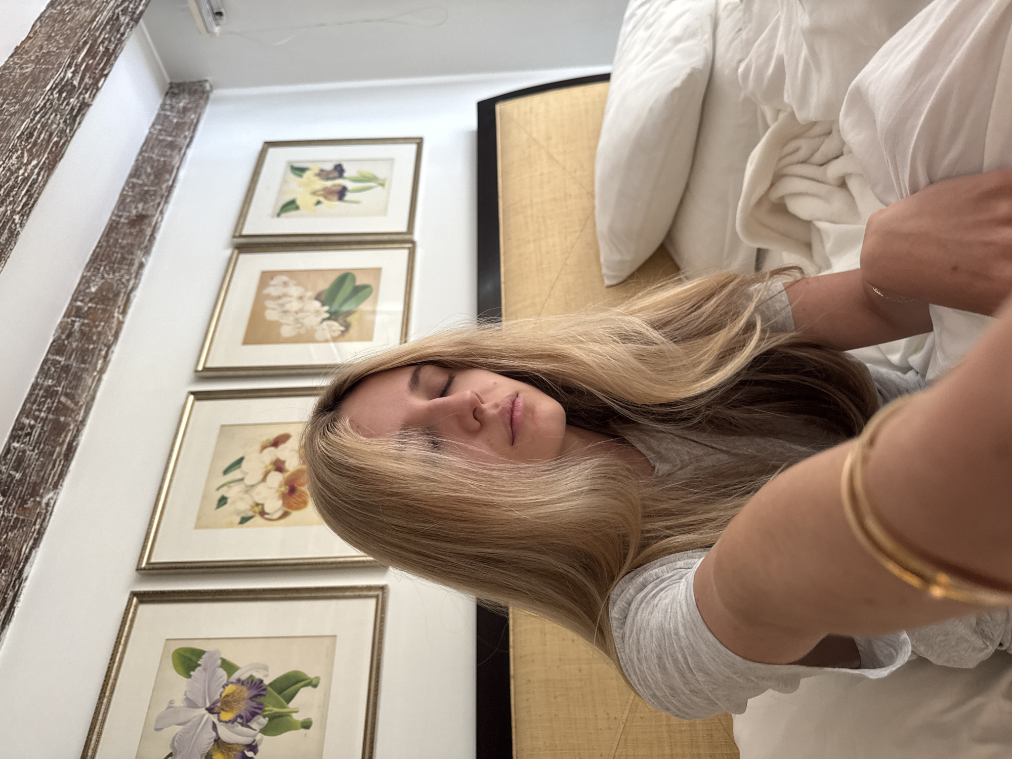 The image is a close-up, selfie-style shot of a young Caucasian woman with long, blonde hair sitting in a bed with white sheets. Her eyes are closed, and she is wearing a grey t-shirt. Behind her, there is a headboard with a woven texture and four framed botanical prints of flowers on the wall. The room has exposed wooden beams on the ceiling. The overall impression is of a bedroom environment. The image does not provide any explicit information or clues about the location being in Ciudad de México, Mexico.
