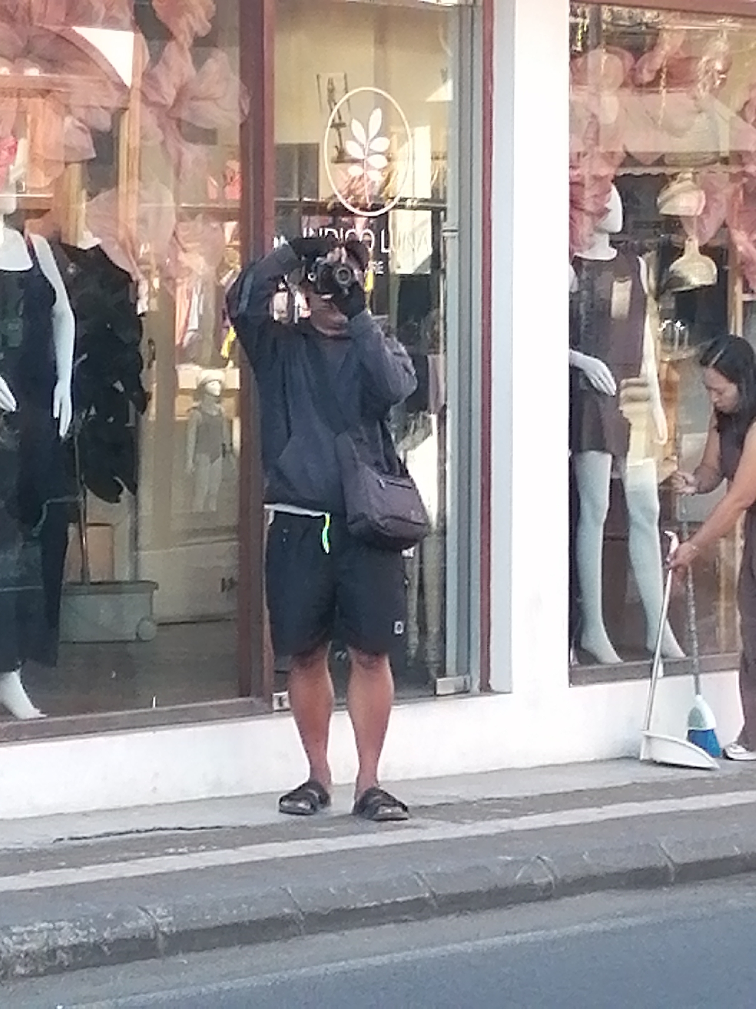 A person is standing on a sidewalk in front of a storefront in Ubud District, Indonesia. The person is holding a camera up to their eye, appearing to take a photo. They are wearing dark clothing, including a jacket and shorts, and sandals. In the background, there are mannequins displayed in the storefront window, wearing clothing. A woman is visible to the right of the frame, holding a broom and dustpan, suggesting she might be cleaning the area. The storefront has large windows and a white pillar on the right side. The sidewalk appears to be made of concrete, and there is a street in the foreground.