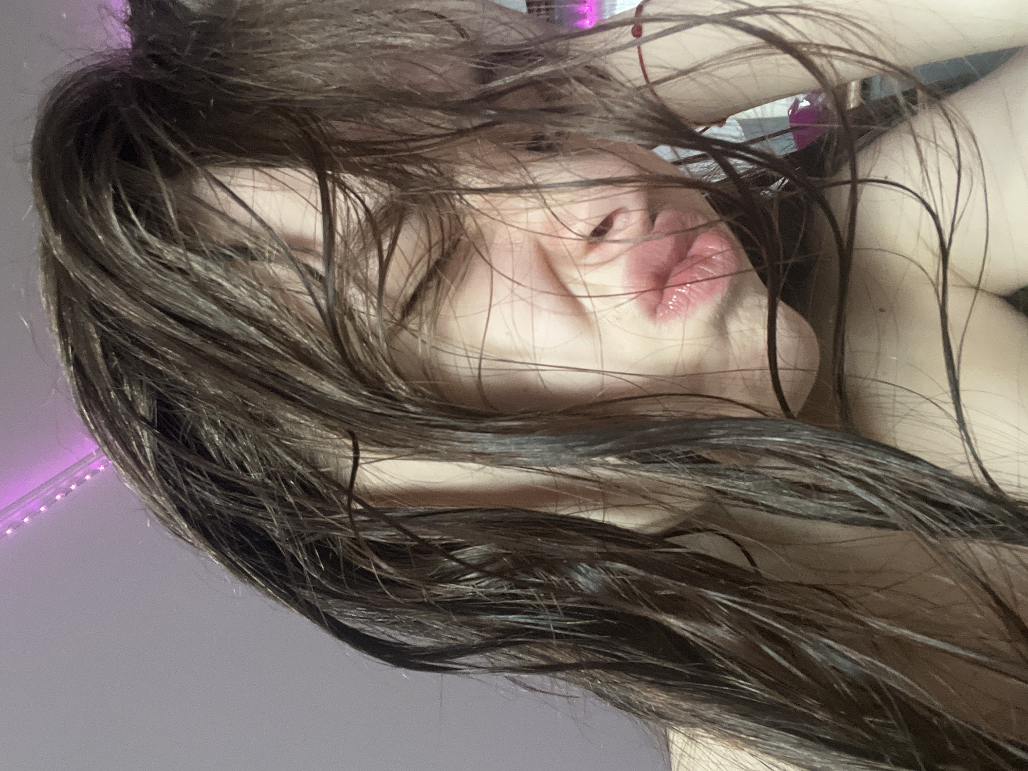 A close-up, slightly rotated image depicts a person with dark, wet hair obscuring much of their face. The focus is on the person's lips, which are slightly pursed. Parts of their skin are visible, appearing pale. In the background, blurred details suggest an interior setting with a hint of pink or purple light, possibly from LED lights.