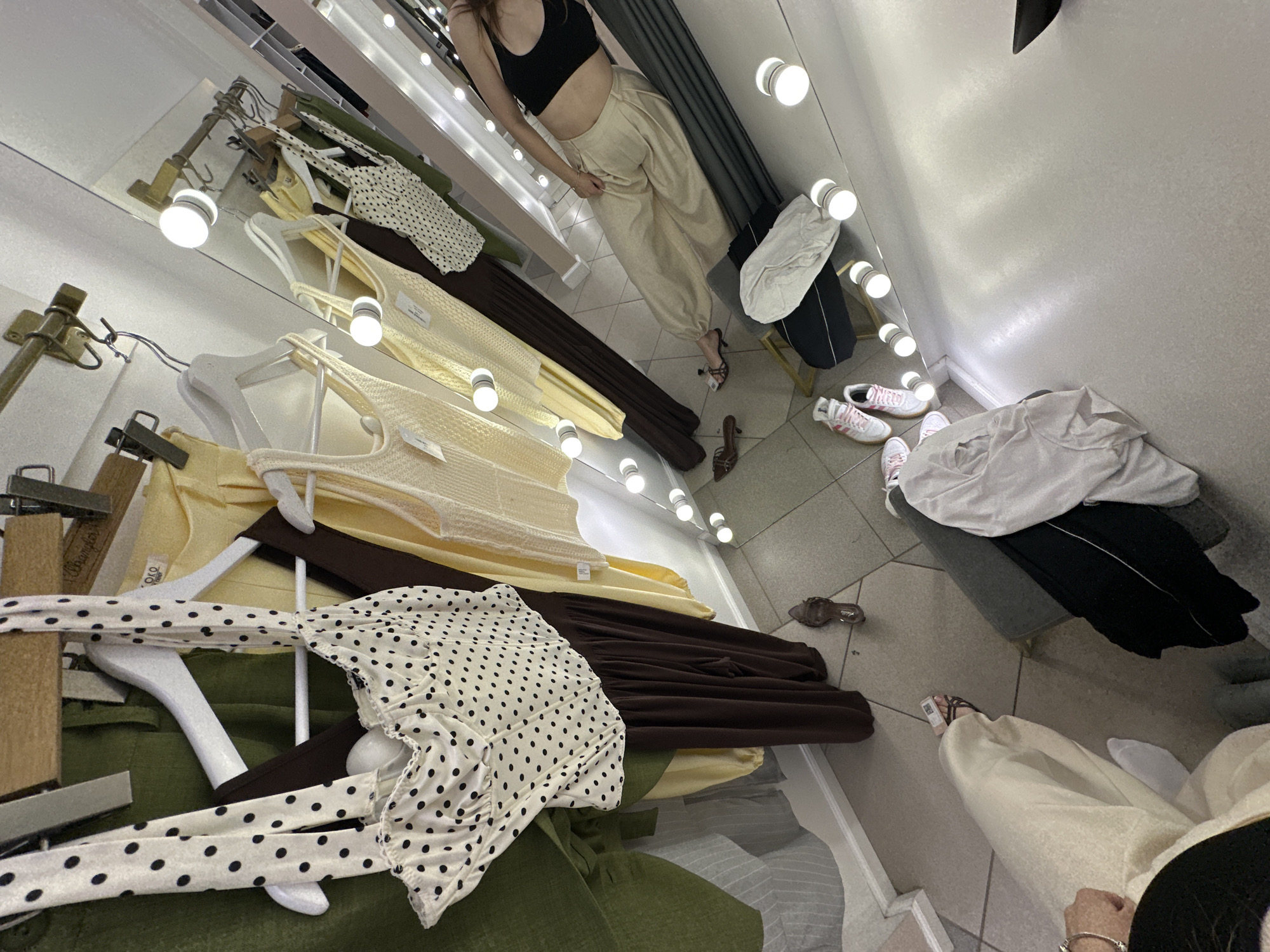 The image shows a dressing room in a clothing store in Perm, Russia. The scene is viewed at an angle, reflecting in a mirror with vanity lights. In the mirror's reflection, a woman wearing a black crop top and beige pants is visible. Garments hang on racks, including a polka dot top, yellow tops, a brown dress, and a green linen shirt. Shoes and sneakers are scattered on the floor. A woman's hand with a bracelet and a small portion of clothing are in the lower right foreground, suggesting someone is in the process of trying on clothes.