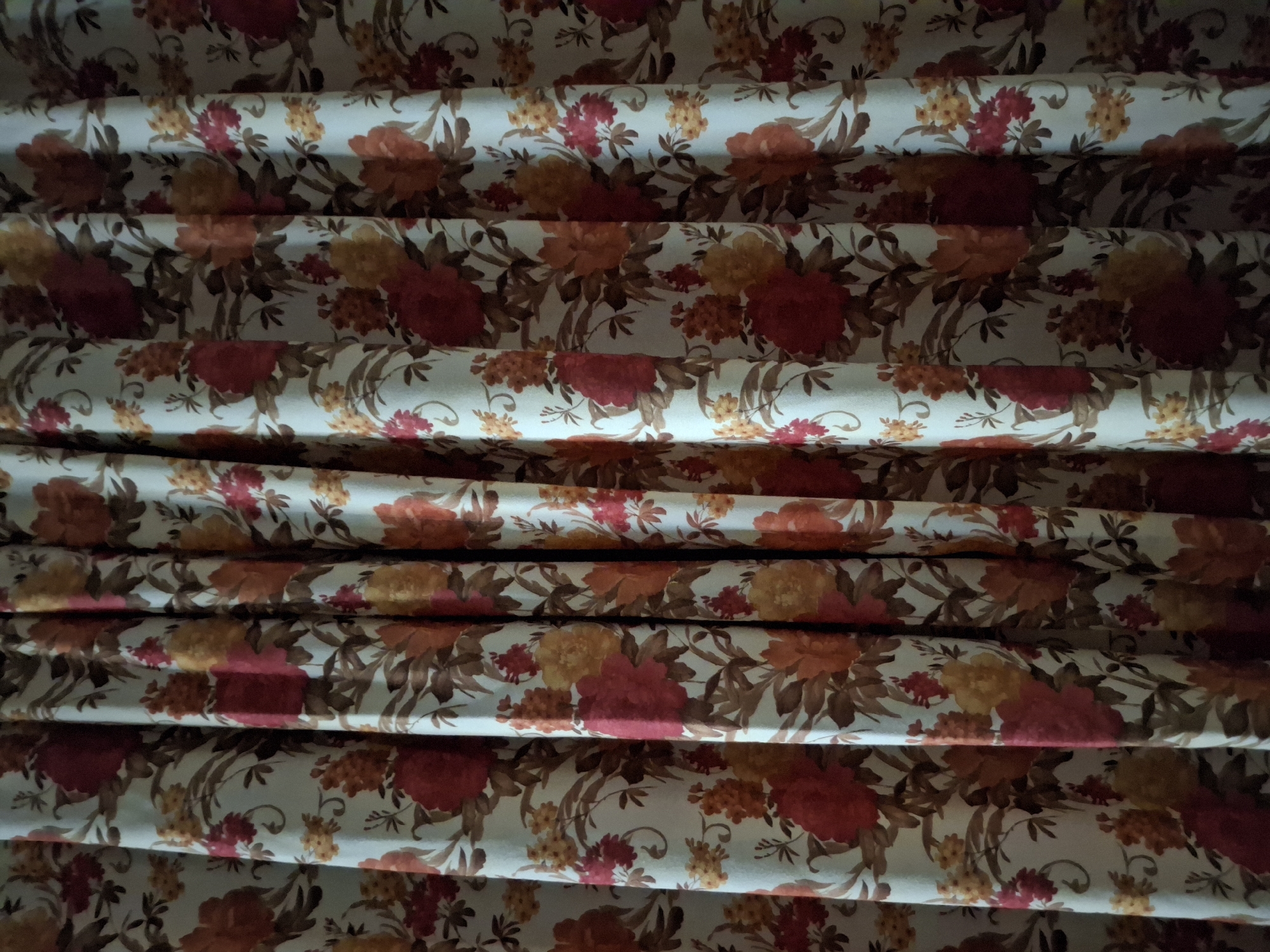 The image shows a close-up of folded fabric. The fabric has a floral pattern featuring red, orange, and yellow flowers with brown leaves against a light background. The folds create a repeating horizontal pattern. The image provides no information regarding the location in Krumovgrad, Bulgaria or any people, subjects, objects, actions, interactions, or environment beyond the fabric itself.