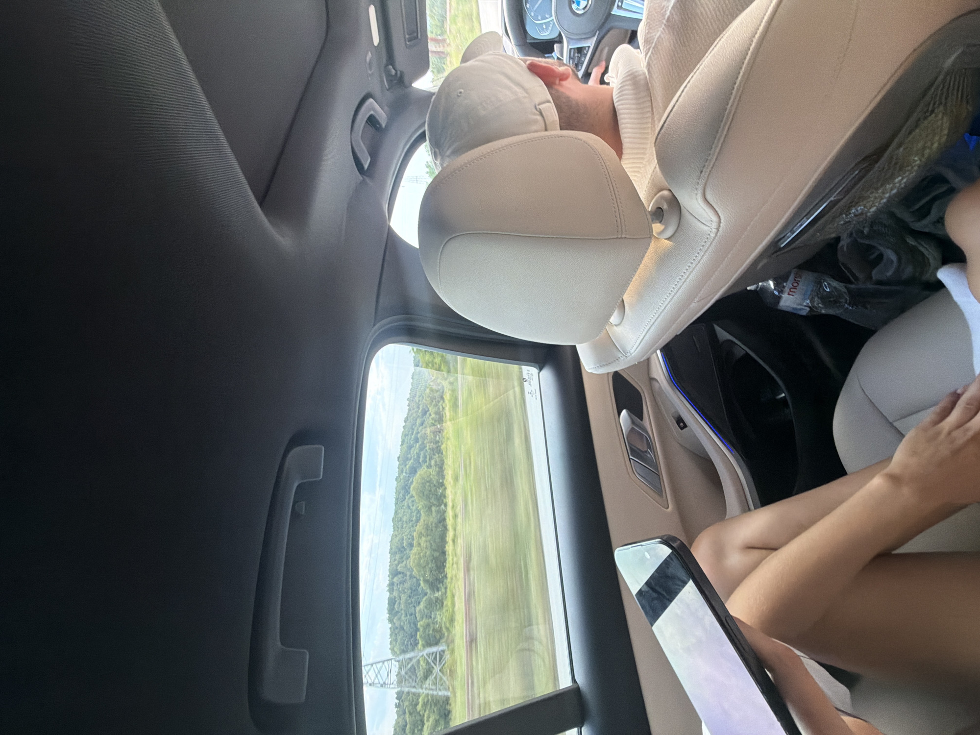 An interior shot from the back seat of a moving car reveals a person wearing a white baseball cap and a cream-colored sweater facing forward. The person's head is partially obscured by a cream-colored headrest. In the foreground, to the left, a person's legs are visible, and they are holding a dark-colored tablet device. The car's interior features light-colored leather seats and ambient blue lighting along the door panel. Through the rear window, a blurred landscape of green trees, a field, and power lines is visible, indicating the car is traveling outside of Onești, Romania, likely on a rural road given the scenery. A water bottle is seen on the seat behind the driver.