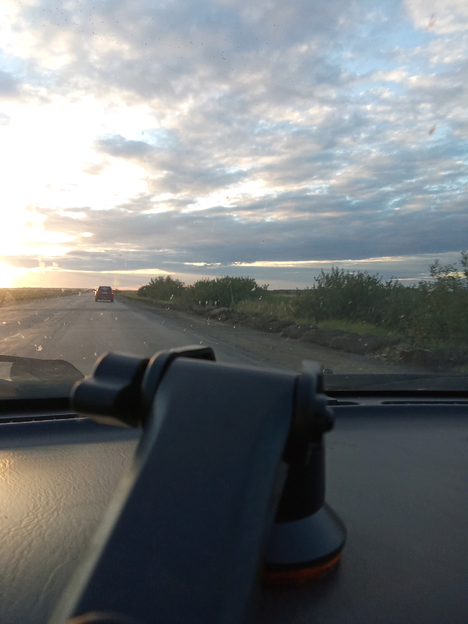 The image is a first-person perspective from inside a vehicle, looking out through the windshield at a rural road during sunset. The sky is filled with a mix of white and gray clouds, with the sun setting on the left horizon, casting a warm, golden light. A dirt road stretches into the distance, with a red car ahead of the viewpoint. Dense green foliage lines the sides of the road. In the foreground, a black car mount for a device is visible on the dashboard. The scene suggests travel through the Светлопольский ауылдық округі in Kazakhstan.