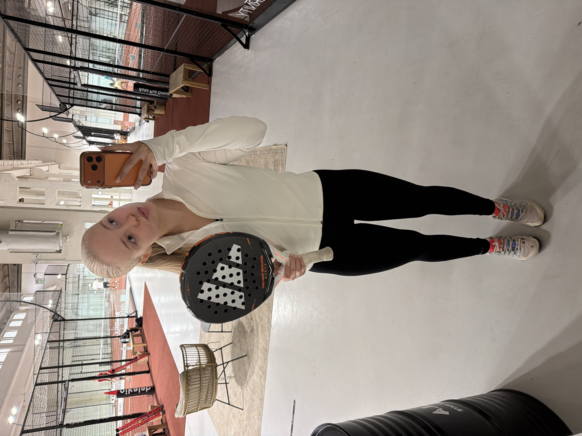A woman takes a selfie indoors at a padel court. She is wearing black leggings, a white top, and athletic shoes. She holds a padel racket in her left hand and an orange iPhone in her right. The background reveals the court with netting and seating, and the lighting is bright. The location appears to be in a sports facility.
