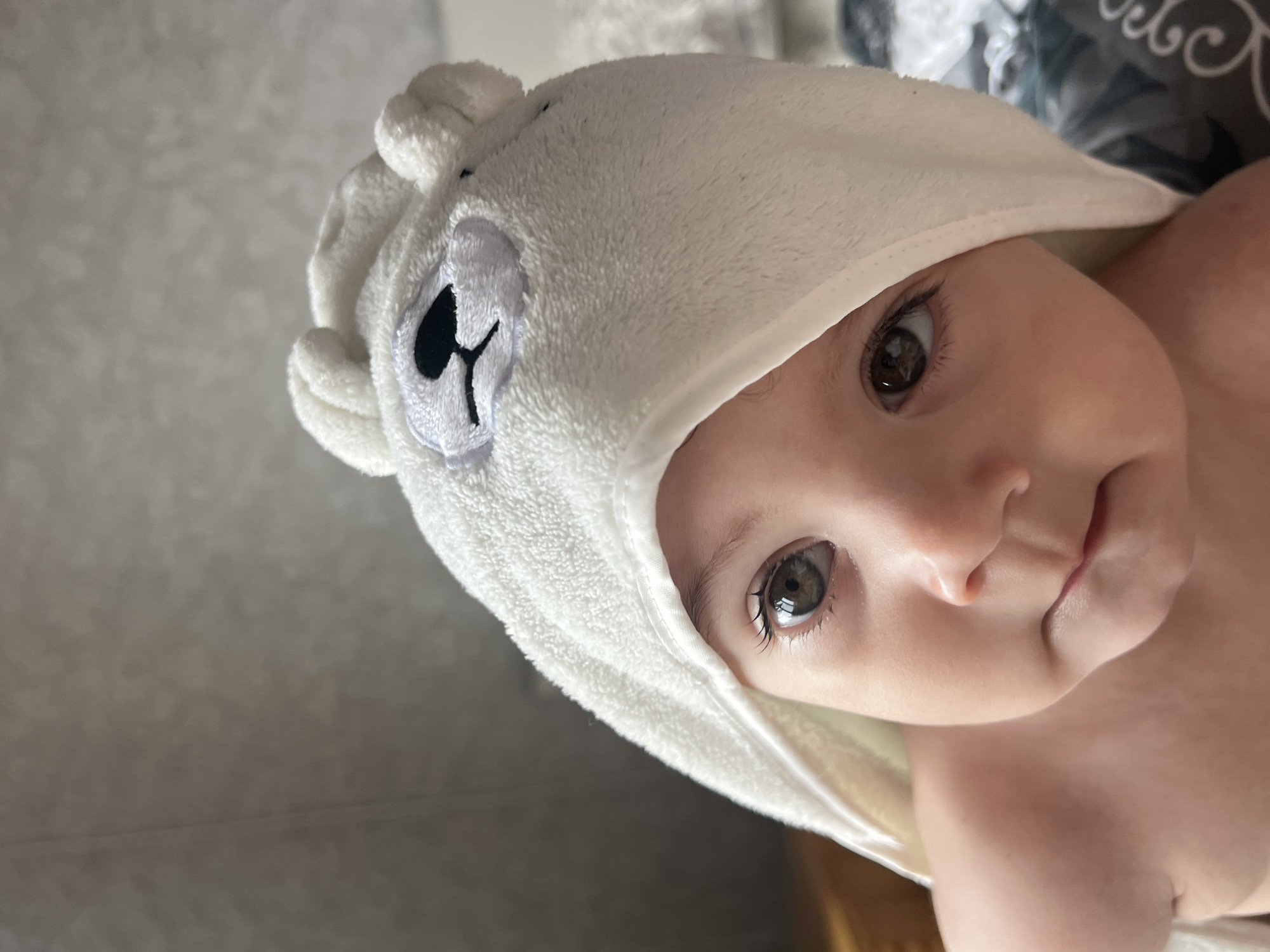 A close-up, slightly angled shot shows a baby wearing a white, hooded towel with a bear design. The baby's face is turned towards the camera, revealing light skin, brown eyes, and a small nose. The bear design on the towel features a stitched face with black details. The background is out of focus, but appears to be a light-colored wall and a partially visible patterned object. There are no identifiable landmarks or features to suggest a location in Elena, Bulgaria.