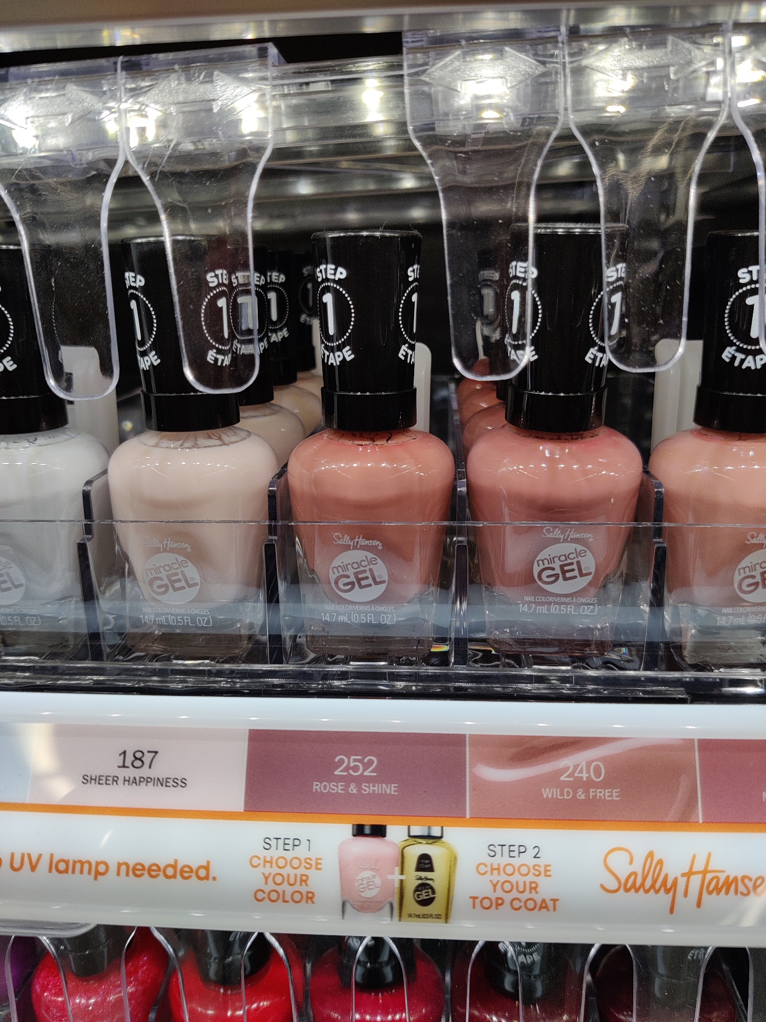 The image shows a retail display of Sally Hansen Miracle Gel nail polish bottles, neatly arranged in clear plastic compartments. The bottles have black caps with white "STEP 1" labels and are filled with various shades of polish, including white, pale pink, and peach. Below the bottles, a shelf displays color swatches with corresponding shade numbers and names: "187 SHEER HAPPINESS," "252 ROSE & SHINE," and "240 WILD & FREE." Prominently featured is text indicating "NO UV lamp needed." The overall environment suggests a well-lit retail store or salon. The location is identified as Mechanicsburg, United States of America.