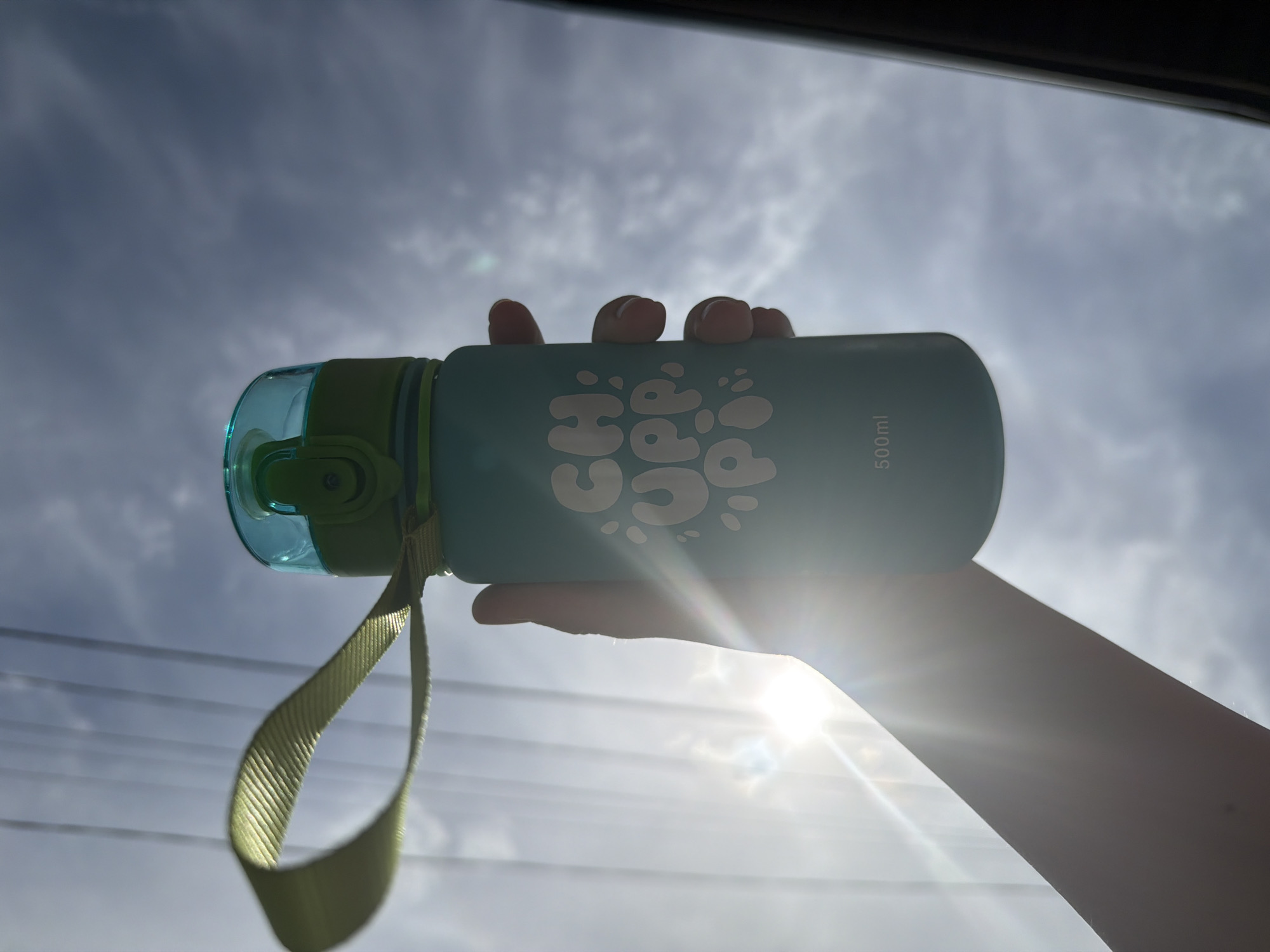 The image shows a hand holding a turquoise water bottle with a light green lid and a yellow strap against a cloudy sky. The bottle has the word "CHUP" printed on it in white letters. The bottle is marked with "500ml". The sun is shining brightly behind the hand and the water bottle, creating a strong glare. The location appears to be outdoors, based on the sky and sunlight. There is a dark object, possibly a car roof or window frame, visible in the upper portion of the image. There is no visible evidence to locate the scene in the city of Chaikovsky, Russia.