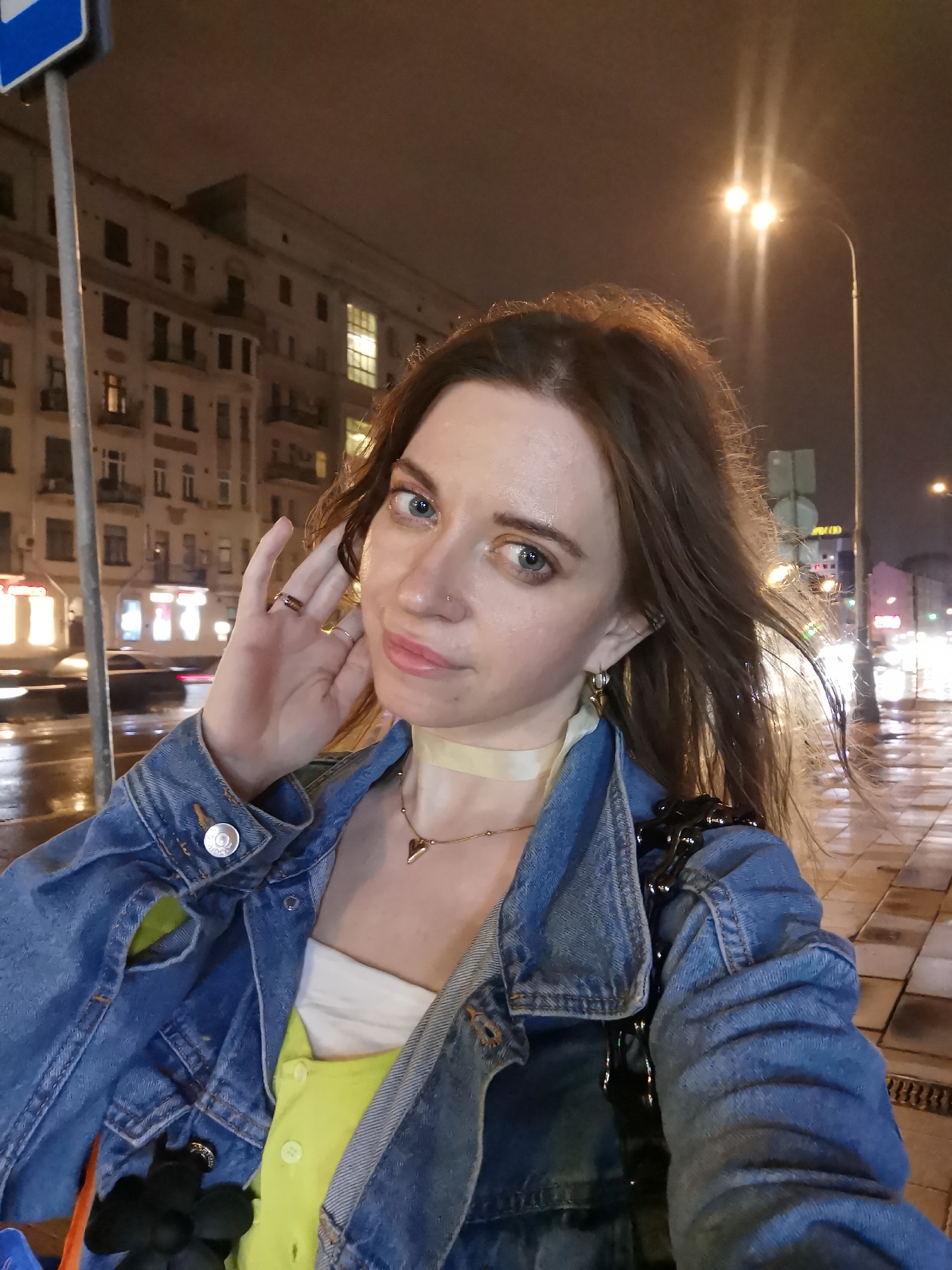 A woman stands on a wet sidewalk at night in an urban environment. She is wearing a denim jacket over a white top and a neon yellow garment, accessorized with a pale yellow choker and a gold necklace with a heart pendant. The woman has brown hair and blue eyes, a nose ring, and is touching her ear with her hand. Several rings are visible on her fingers. In the background, a multi-story building is illuminated by lights, and streetlights line the wet street. Blurred car lights and other signage indicate vehicular traffic. The location is identified as a city of Unknown, Unknown.