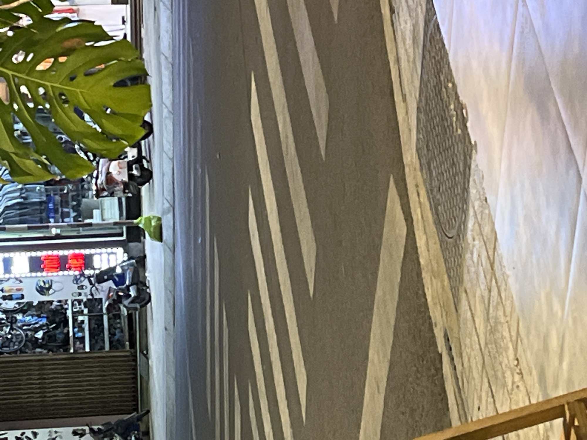 The image captures a street scene in Nha Trang Ward, Vietnam, focusing on a pedestrian crossing. In the background, a shop displays various items, with a motorcycle parked in front. A large, green leaf from a Monstera plant hangs into the frame from the top right, partially obscuring the view of the shop and a second motorcycle. The pedestrian crossing is clearly marked with white stripes on the asphalt road. The foreground shows a sidewalk with tiled paving and a manhole cover. The lighting suggests it is either early morning or late evening, with artificial lights illuminating the shop.