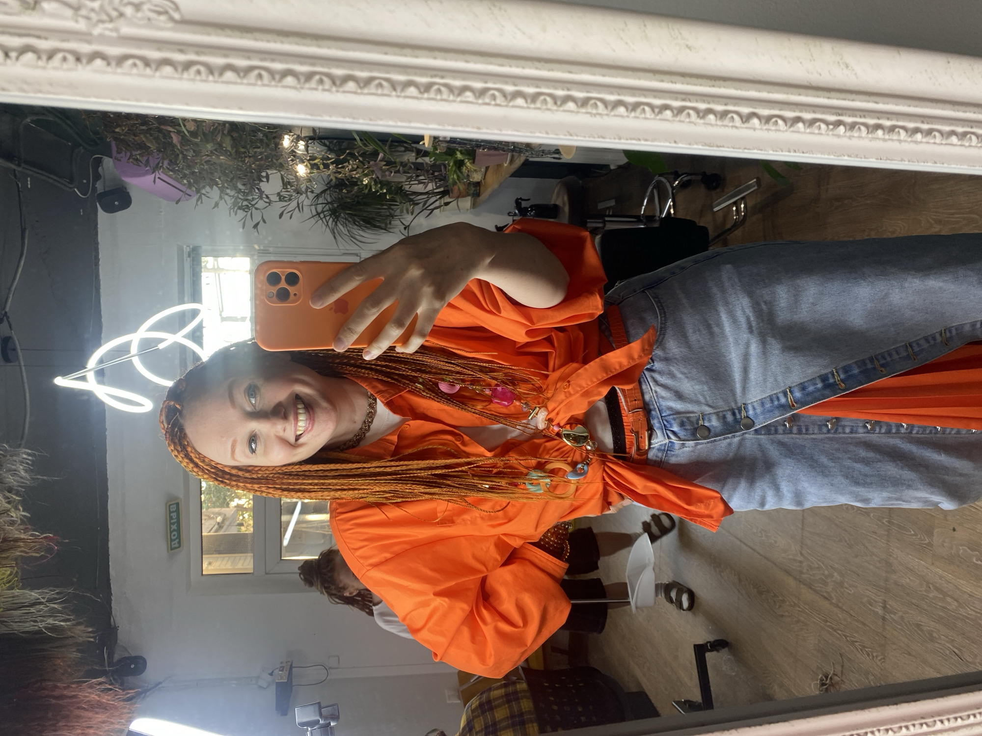 A mirror selfie captures a smiling woman with long, vibrant orange box braids, reflecting her image and the interior of what appears to be a hair salon or creative studio in Sochi, Russia. She holds an orange-cased smartphone in her right hand, taking the photo.
The woman is wearing an orange collared shirt or jacket tied at the waist, layered over a white top. She has several decorative necklaces with colorful charms and a thicker gold-toned chain around her neck. Her lower body is clad in a light blue denim garment, possibly a skirt or dress, with buttons down the front and a brown belt visible.
The reflection reveals a bright indoor space. In the background, lush green and reddish artificial or real plants are arranged. A white, swirling neon light fixture is prominent on the wall behind her head. To the left, a portion of another person with dreadlocks is visible. Further back, a window shows natural light coming in, suggesting it is daytime. Above a doorway in the reflection, the green Cyrillic text "ВЫХОД" (Exit) is visible.
Various pieces of equipment, including what looks like a ring light and metal stands, are reflected on the floor. The floor itself is made of light-colored wood. The mirror has an ornate, white decorative frame across the top. The woman's broad smile suggests a cheerful and positive mood.