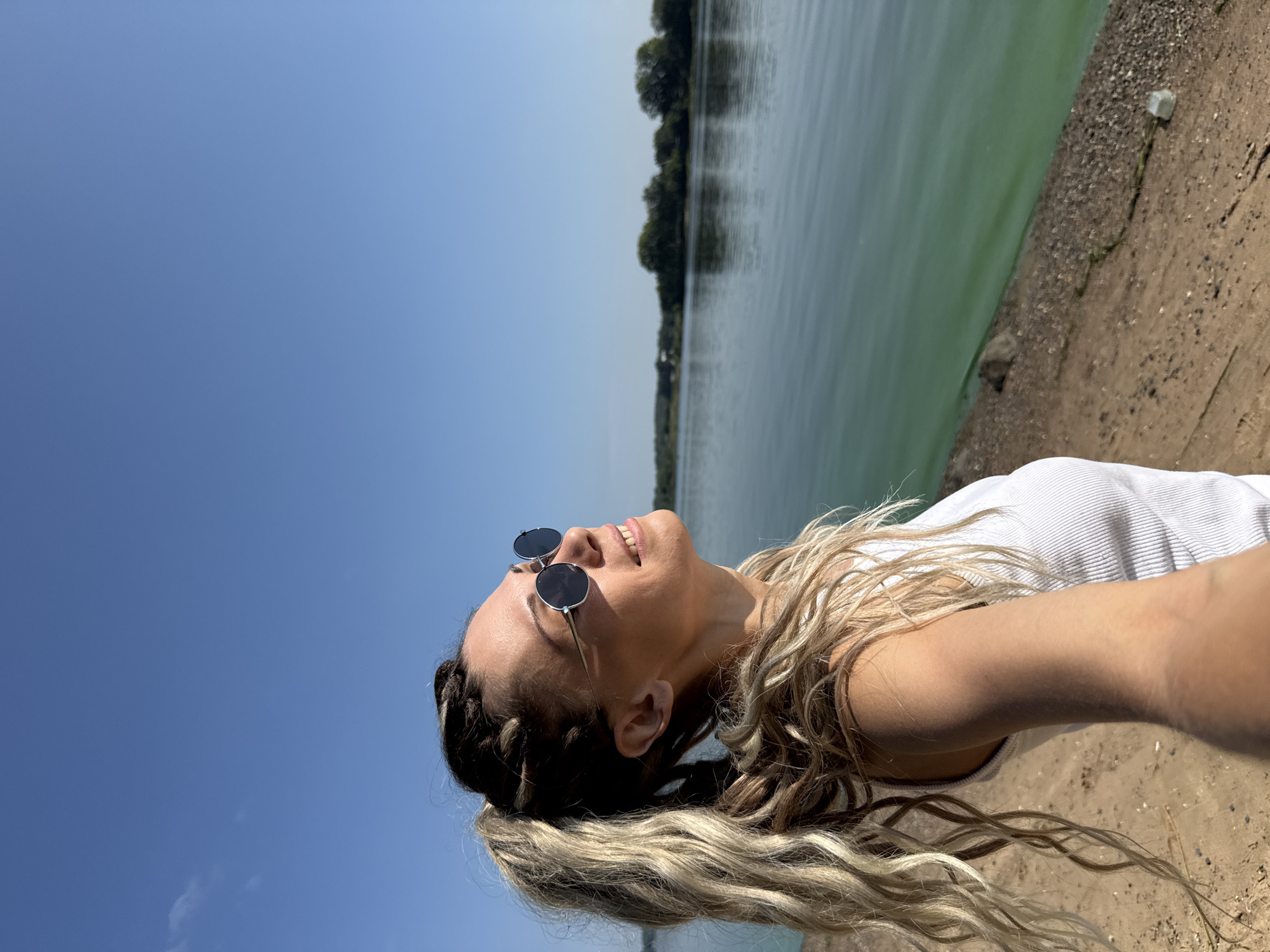 A medium shot captures a woman with blonde dreadlocks styled in a high ponytail, facing left, with her head tilted upward toward the sky. She is wearing round, dark-lensed sunglasses and a white ribbed tank top. The background features a body of green water with reflections of trees on the opposite bank, under a clear blue sky. The scene appears to be outdoors, possibly near a waterfront.