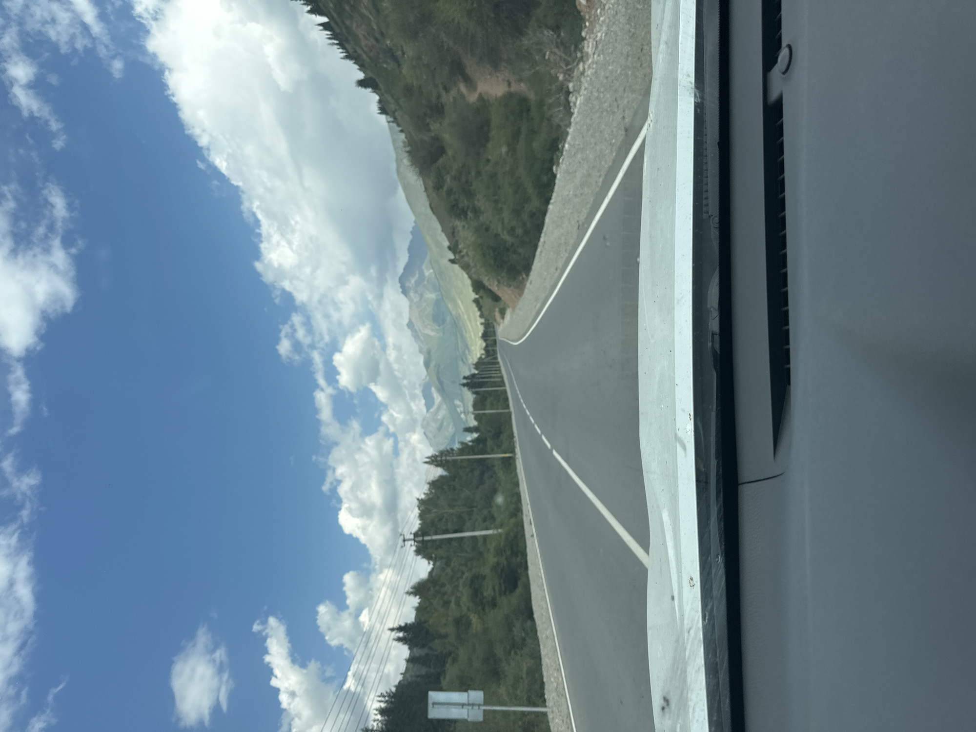 The image is a first-person perspective from inside a vehicle, looking out at a scenic road winding through a mountainous landscape. The road is paved with a white dashed line marking the center and a white solid line on the right edge. Power lines and poles run along the left side of the road, with dense green trees and bushes on either side. In the distance, majestic mountains rise under a bright blue sky dotted with white clouds. The foreground shows the dashboard of the vehicle, partially obstructing the view. The scene appears to be a highway or a main road in a rural or mountainous area, possibly near Semenovka, Kyrgyzstan, given the characteristic terrain and vegetation. There are no people or active events visible in the image.