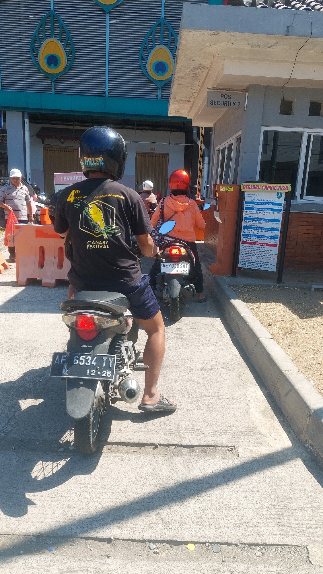 The image depicts a scene at a security post with a toll gate, likely in Nologaten, Indonesia, given the presence of a sign indicating "BERLAKU 1 APRIL 2026" and "PASAR LEGI PONDOROSO" which translates to "POS Security 2" and "Valid Until April 1, 2026" and "Pondoroso Legi Market" respectively. In the foreground, a person wearing a black helmet and a black t-shirt with a "CANARY FESTIVAL" graphic is seated on a motorcycle. The motorcycle has a license plate reading "AE 6534 TY" and "12-26". Another person wearing an orange jacket and a red helmet is on a motorcycle behind the first. This second motorcycle has a license plate "AE 4030 SAT" and "06-30". A third person, wearing a white cap and a striped shirt, is visible to the left, near an orange traffic barrier. The environment is outdoors, with a paved walkway and a building in the background. The sky is bright and sunny, casting shadows. The overall scene suggests a routine day at a checkpoint or market entrance.