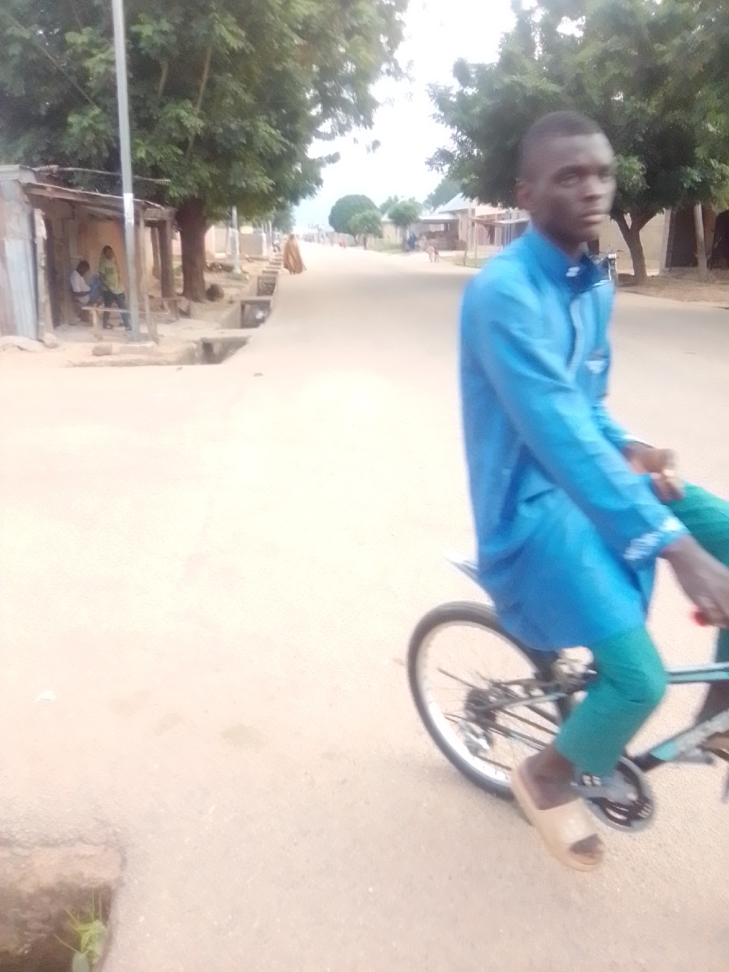 A young Black male wearing a blue long-sleeved shirt and green pants rides a bicycle on a dirt road. The man is positioned in the foreground on the right side of the image, facing left and slightly away from the camera. The bicycle's rear wheel is visible. In the background, on the left side, a small open-air structure with a corrugated metal roof and wooden posts is visible. Two individuals, one wearing a green headscarf, are seated under the structure. A dirt road with scattered debris stretches into the distance, lined with trees and small buildings. The scene appears to be an outdoor environment in a rural or semi-urban area, possibly in Liji Hill, Nigeria, given the cultural attire and the general setting. The lighting suggests it is daytime, with an overcast sky.