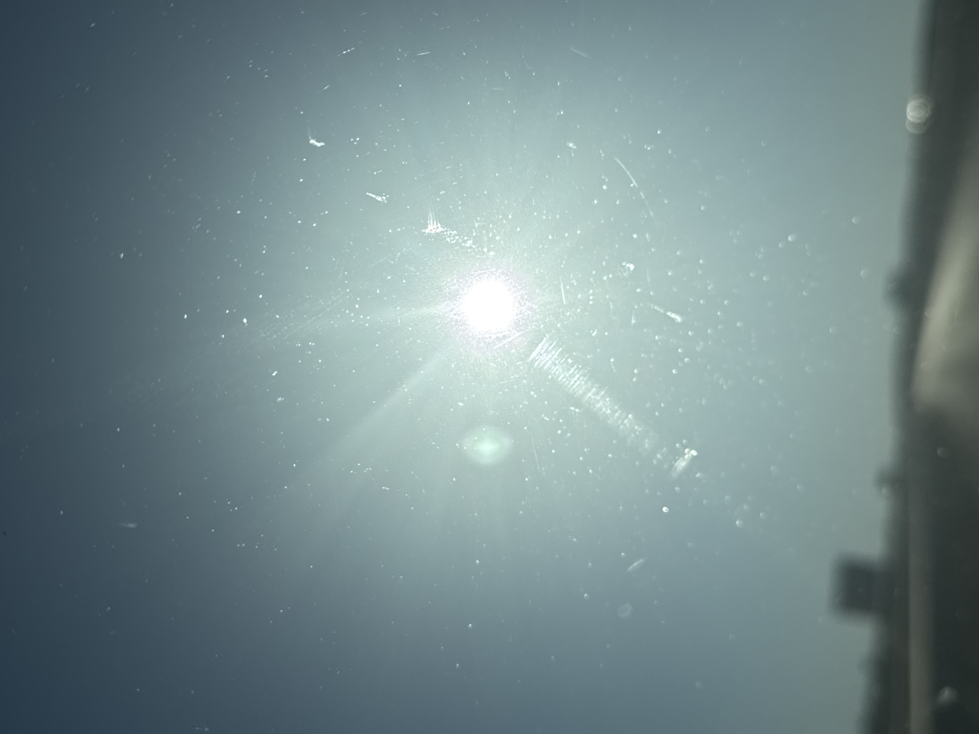 The image is a low-angle shot of the sun, viewed through what appears to be a windshield. The sun is the brightest point in the image, radiating light outwards and creating lens flare effects. Numerous small white specks are visible, likely dust or water droplets on the glass. The background is a hazy, light blue sky. In the lower portion of the image, a blurred road and a dark, indistinct object that may be a vehicle or a building are visible. The image captures a moment of bright sunlight, possibly during travel. There is no discernible indication of the specific city of Vratitsa, Bulgaria.