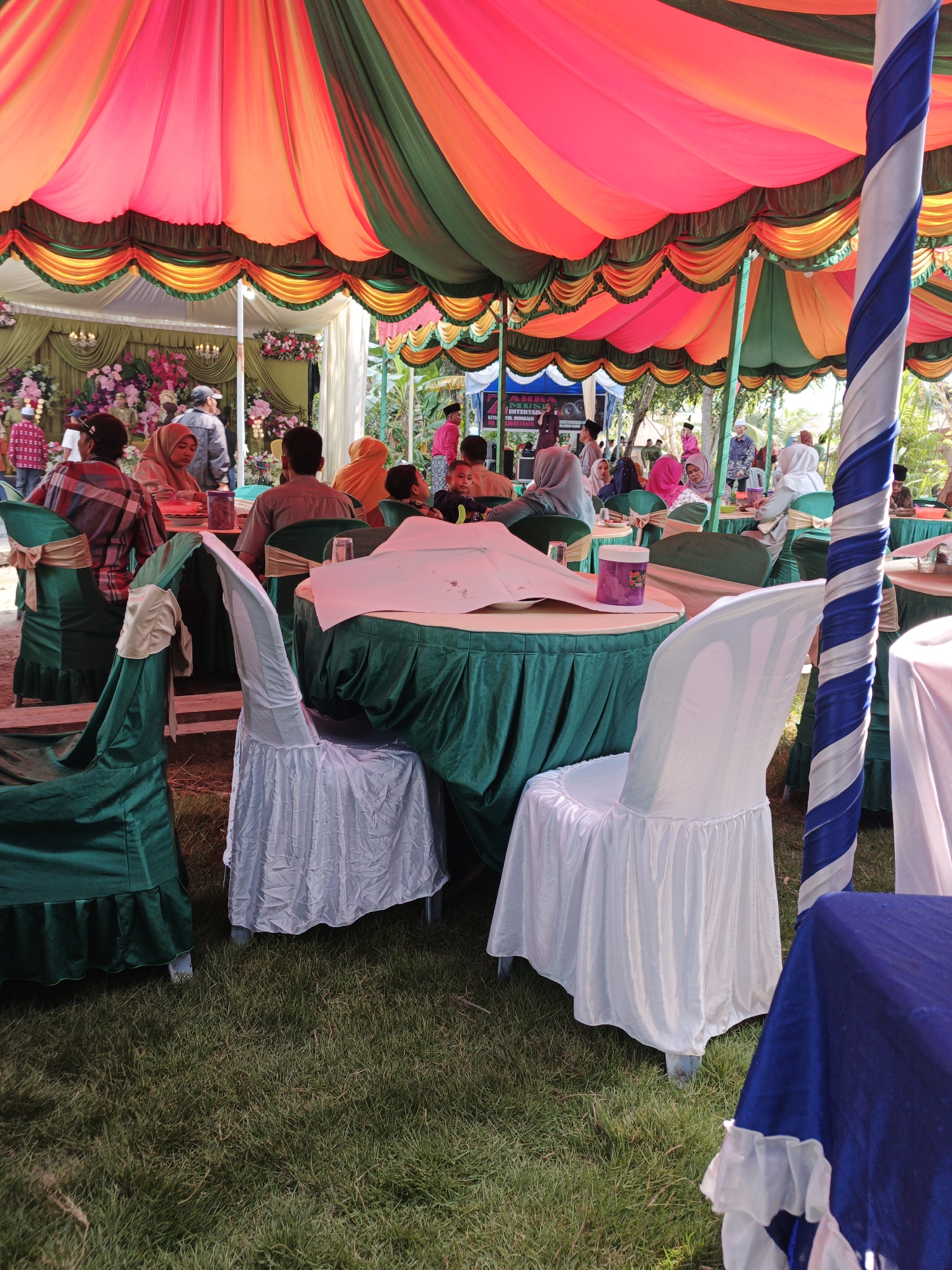 An outdoor event is underway under a colorful striped tent with pink, orange, and green fabric. Several round tables are covered with green tablecloths and decorated with draped fabric on the chairs. Some chairs are covered in green fabric with gold sashes, while others are covered in white fabric. In the foreground, a table with a blue tablecloth is visible. People are seated at the tables, and others are standing or walking around. Some individuals are dressed in traditional Indonesian attire, while others are in casual clothing. The event appears to be a gathering or celebration, possibly a wedding or religious festival. The ground is covered with grass, and trees and foliage are visible in the background, suggesting a rural or semi-rural setting. The overall impression is one of a festive and communal occasion. The presence of traditional decor and attire suggests this event is taking place in Pematang Duku, Indonesia.