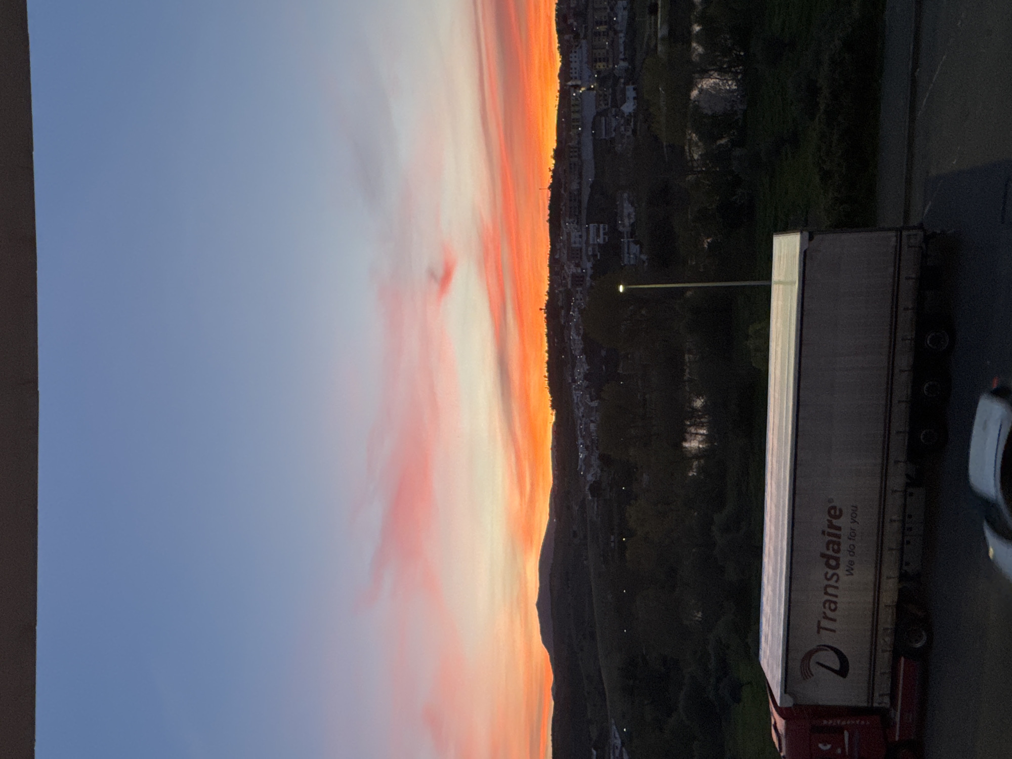 The image shows a street scene, likely in the city of Mirandela, Portugal, based on the architecture in the background. A large "Transdaire" truck is parked to the right of the frame. The sky on the left is a gradient of blue to pink and orange, suggesting either sunrise or sunset. A line of buildings can be seen along a hill, with vegetation also visible. A streetlight is in the middle of the image, partially obscured by trees. A partial view of a car is visible on the bottom right.