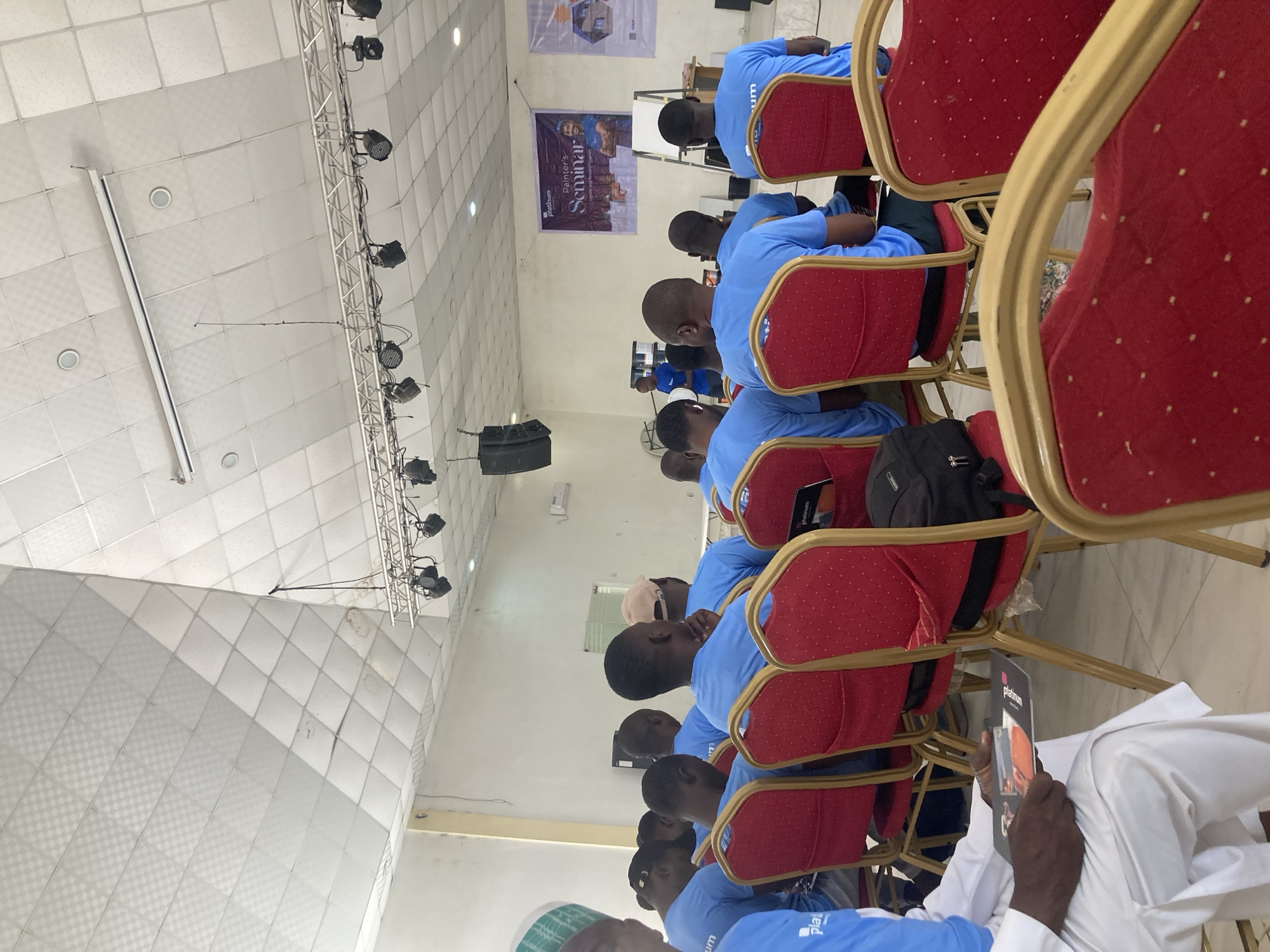 The image shows a group of people sitting in an auditorium. They are seated in red chairs with gold frames and are mostly wearing light blue shirts. The location is possibly in Jos, Nigeria. The people are attending what appears to be a seminar, as indicated by a banner in the background that reads "Platinum Painter's Seminar." There is also stage lighting attached to the ceiling. A black backpack is visible hanging on a chair. In the foreground, a person wearing white clothing is holding a brochure with the word "Platinum" on it.