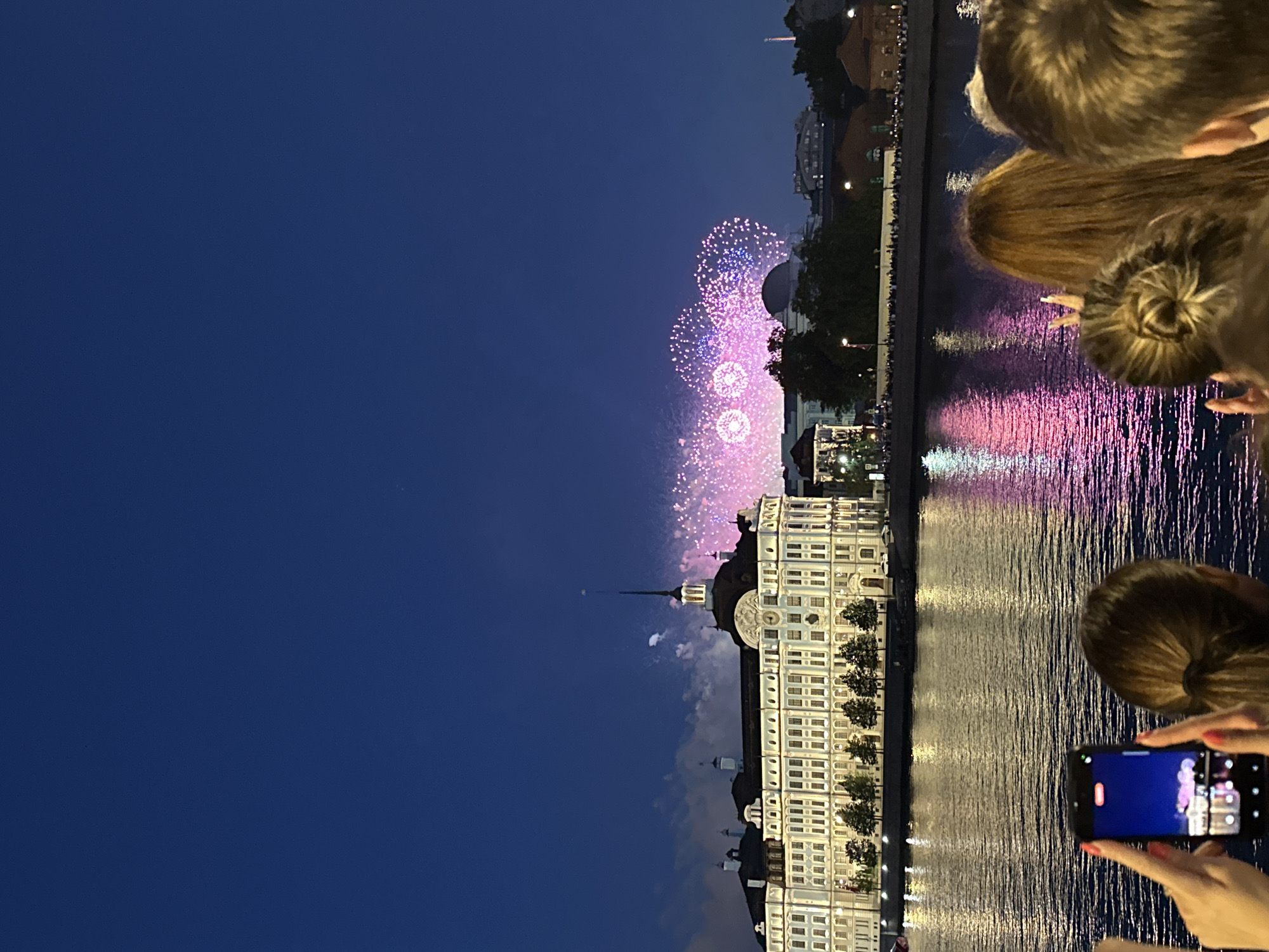 This image captures a twilight view of the Neva River in Saint Petersburg, Russia, during a fireworks display. In the background, a grand, illuminated building, likely a historical palace or administrative center, stretches across the horizon. Its windows are lit, and the facade is ornate. The river in the foreground reflects the colorful fireworks and the building's lights, creating a shimmering spectacle. Several fireworks bursts, primarily in shades of pink and purple, illuminate the dark blue sky above the cityscape. In the immediate foreground, the backs of several people are visible, indicating they are spectators watching the fireworks. One person on the left is holding up a smartphone, capturing the event. The overall scene suggests a festive evening event.