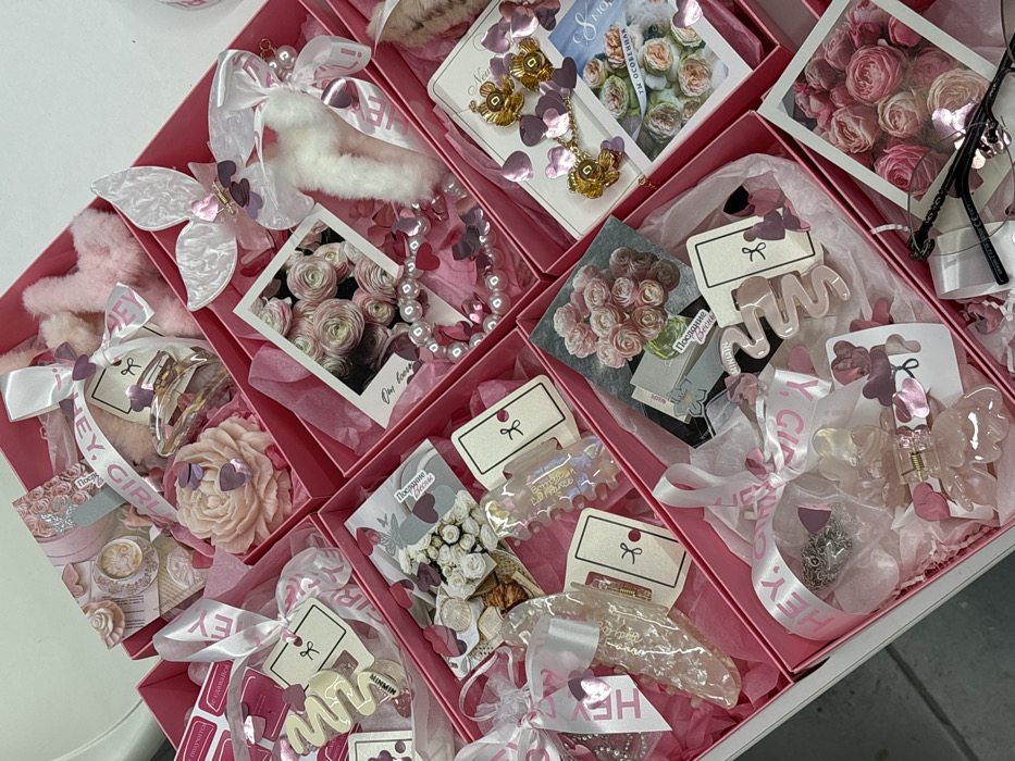 The image displays an overhead, slightly angled view of multiple open, square, pink cardboard gift boxes, likely arranged within a larger segmented container, resting on a light-colored reflective surface. Each box is lined with pink tissue paper or shredded material and contains a distinct assortment of feminine-themed items, predominantly in shades of pink, white, and gold.
Contents include various plastic hair claw clips in designs such as an 'M'-shape, a wavy form, a floral shape, a butterfly, and a mermaid tail adorned with a white faux fur pompom. Also visible are a large pink rose-shaped candle or soap, a pearl-style bracelet with pink heart charms, gold-toned dangle earrings featuring floral elements and pink heart-shaped sequins, and black-framed eyeglasses with a pink charm. Additional items consist of small pink furry accessories, clear iridescent decorative bottles, and numerous small prints or cards depicting arrangements of pink roses, one in a polaroid style, and another showing a coffee cup. White satin ribbons with "HEY, GIRL!" printed in pink are tied around several items. Scattered pink and purple heart-shaped confetti are distributed throughout the boxes. Several tags and cards bear Russian text, including "Послание Весны" (Message of Spring), "С 8 Марта!" (Happy March 8th!), and "Ты особенная" (You are special).
The scene represents an interior workspace or retail packaging area. Based on the presence of Russian text and the prompt's context, the location is within Krasnodar, Russia, although precise street or building identification is not discernible from the visual information. No people are present.