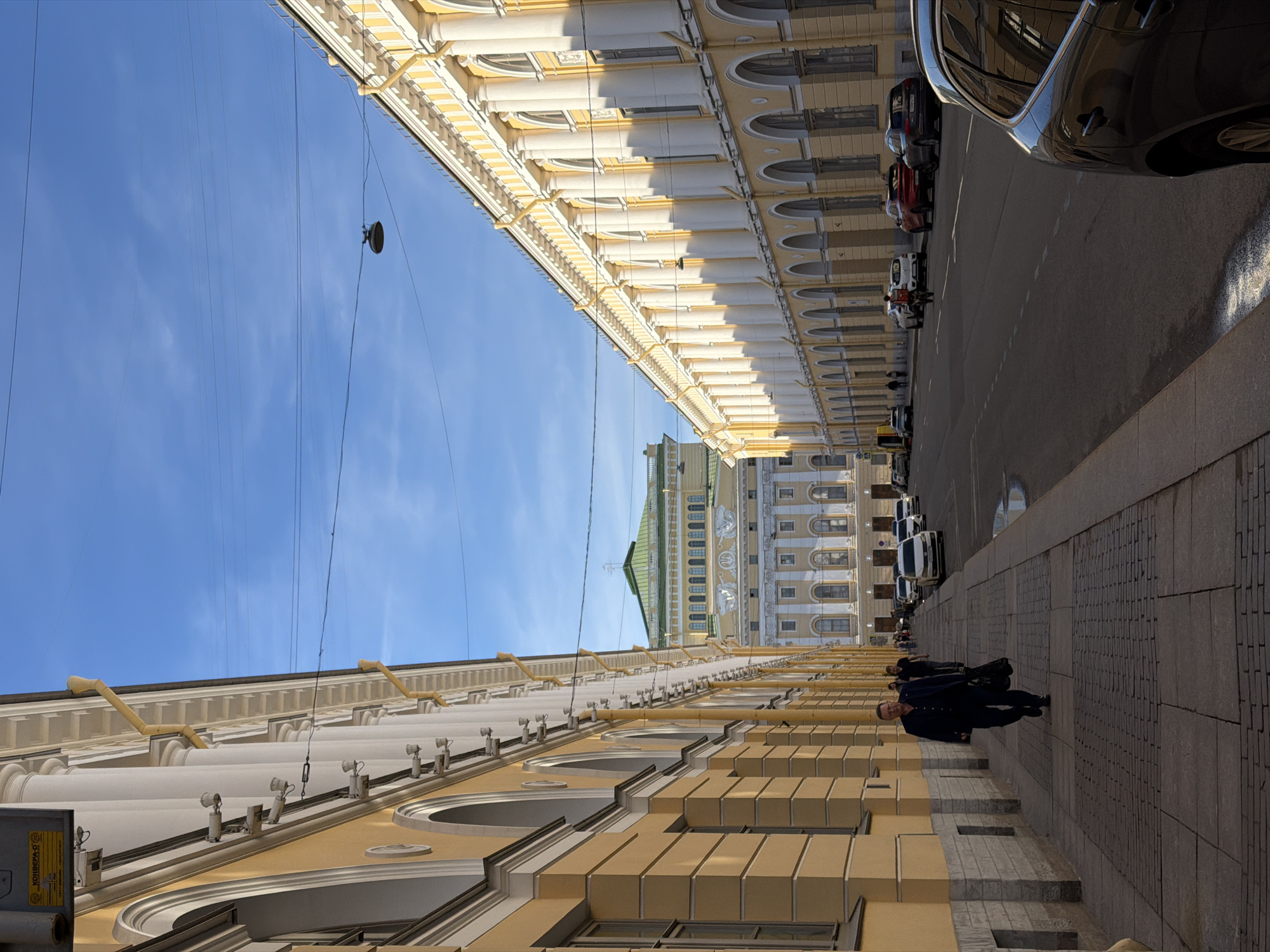 This image captures a street scene in Saint Petersburg, Russia, likely in the historical center given the architecture. The perspective is looking down a wide street lined with grand, yellow neoclassical buildings featuring arcaded ground floors and repetitive rows of windows and columns on upper levels. The sky is a clear, bright blue with some wispy clouds. Overhead, electrical wires and a street lamp are visible.
On the left side of the street, a man in a dark suit walks away from the camera on a paved sidewalk. He is carrying a large black bag. Further down the sidewalk, other pedestrians can be seen walking. On the right side of the street, several cars are parked and some are driving, including a black SUV in the foreground on the right. In the distance, a large, ornate building with a green roof and classical detailing is prominent. The street itself is asphalted, with visible lane markings. The overall impression is one of urban grandeur and a pleasant, sunny day.