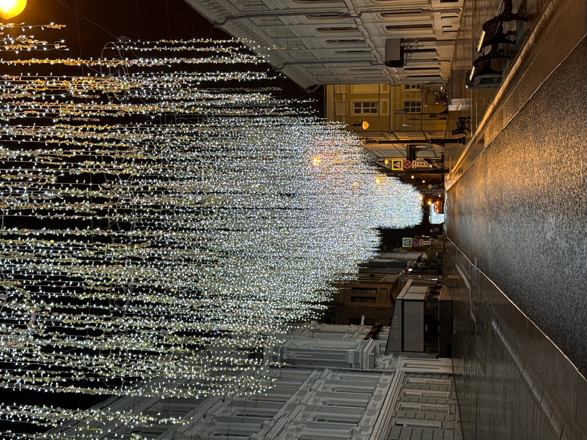 The image depicts a street scene at night in Moscow, Russia, adorned with holiday lighting. The street is lined with buildings, some with ornate facades and illuminated windows. Overhead, strings of small, white lights create a luminous canopy. The street appears wet, reflecting the lights and adding to the ambient glow. Street furniture such as benches are visible along the sidewalk. Traffic signs are also present.