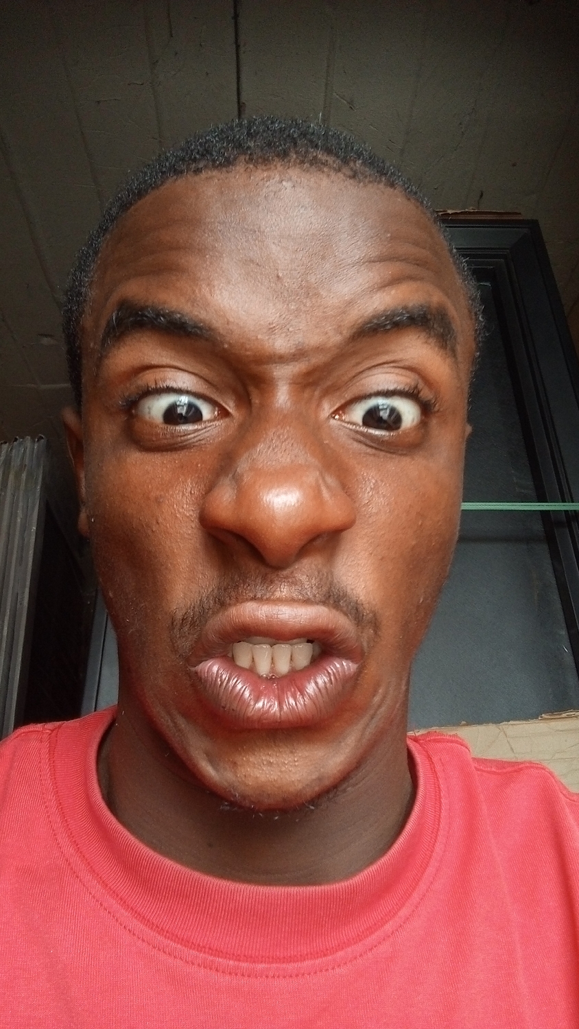 This close-up portrait, likely a selfie, features a young Black male directly facing the camera in Nkpor, Nigeria. He is making an exaggerated and humorous facial expression, characterized by wide-open eyes with visible whites, and a contorted mouth pulled back into a grimace, revealing his upper and lower front teeth. His eyebrows are furrowed, creating prominent wrinkles on his forehead.
The individual has short, dark hair, and subtle facial hair in the form of a light mustache and stubble around his chin and jawline. He is wearing a bright red, crew-neck t-shirt, which occupies the lower portion of the frame.
The background is dimly lit and mostly out of focus, showing dark, indistinct architectural elements, suggesting an indoor or shaded outdoor setting. The lighting appears to be natural, indicating it was taken during the daytime. The overall mood conveyed by the subject's expression is one of playful surprise, shock, or intense focus. No discernible text is visible in the image.
