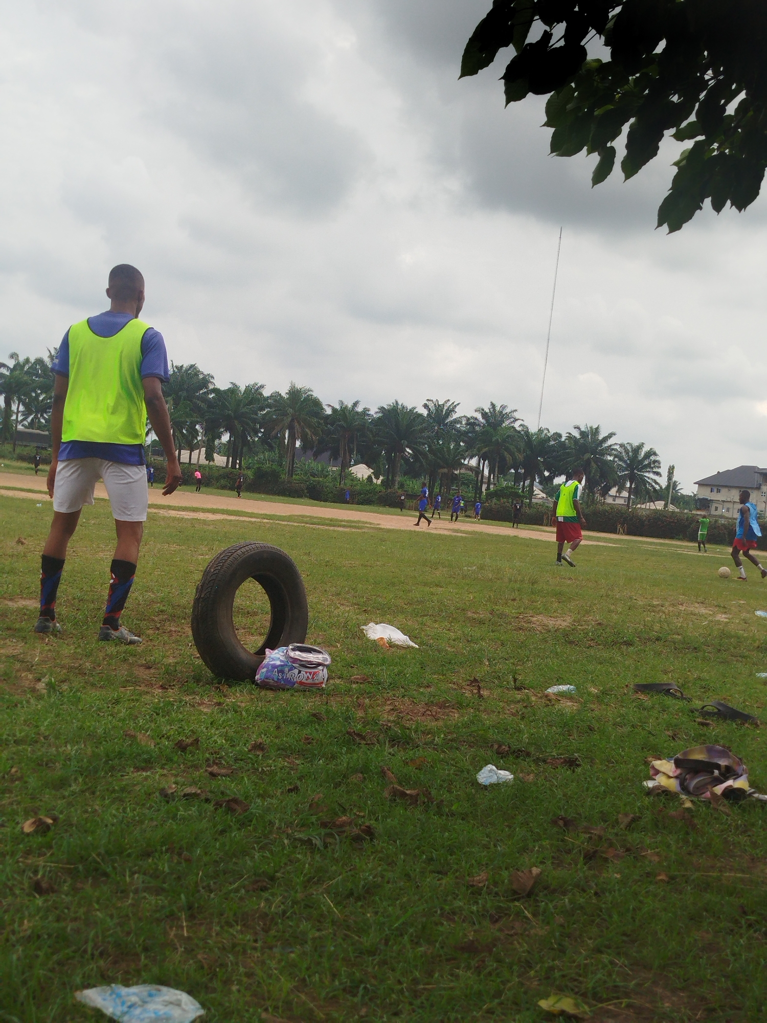 The image depicts a grassy field in Ikot Mbonde, Nigeria, with several individuals engaged in a soccer game in the background. In the foreground, a man wearing a blue shirt, white shorts, and a bright yellow vest stands with his back to the camera. Next to him is a tire and some discarded packaging on the ground. The environment shows lush green vegetation, with palm trees lining the background, and a cloudy sky. The field contains litter scattered across the grass. Several other players wearing soccer gear can be seen participating in the game further away. In the distance, there's a building.