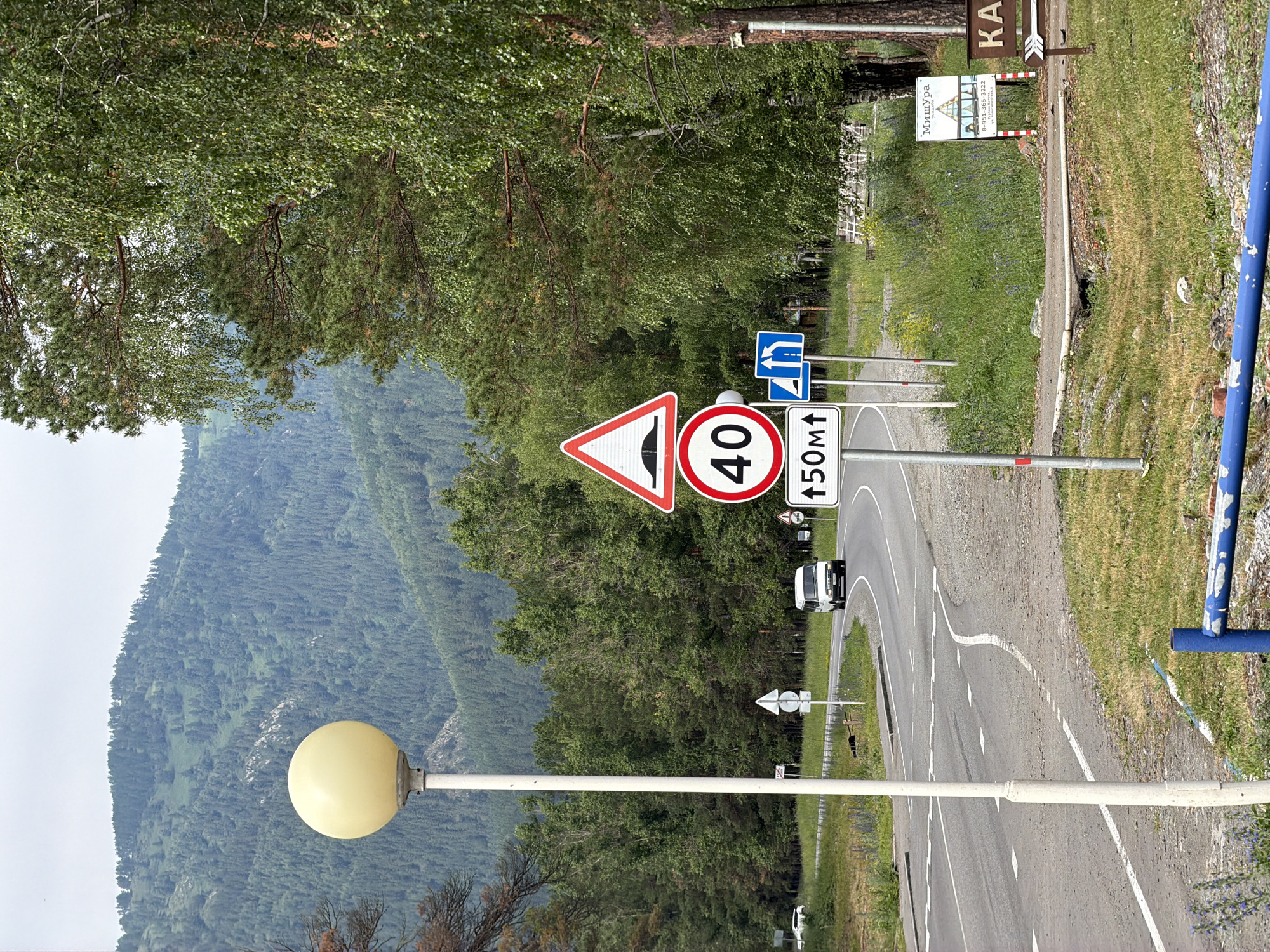 The image depicts a scenic view of a road winding through a lush, green mountainous landscape. The road itself is paved and has white lane markings, with a speed limit sign indicating 40 and a hump or speed bump warning sign visible. Another sign shows an arrow pointing upwards with "50М↑". A white van is driving on the road. Along the side of the road, there is a tall lamppost with a spherical yellow light. The background is dominated by a densely forested mountainside, suggesting a natural, possibly rural or remote setting. The trees are vibrant green, indicating it is likely summer or spring. The sky is visible in the upper left, appearing bright and overcast, suggesting daylight hours. There are also signs indicating directions and possibly business names. One sign has Cyrillic text that reads "Мишура" and a phone number, suggesting a local establishment. Another sign has the letters "КА" prominently displayed. The overall impression is one of a peaceful, natural environment with a road providing access.