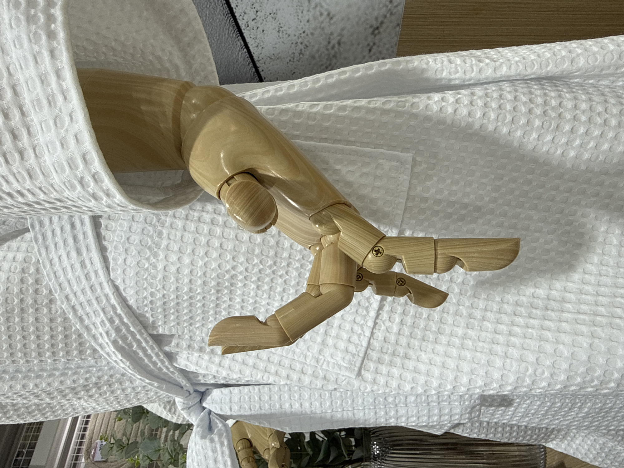The image shows a wooden mannequin arm dressed in a white waffle-weave robe. The arm is positioned in front of the robe, with the hand bent at the wrist and fingers partially extended. The robe is tied with a belt at the waist. In the background, there is a blurred view of greenery and a ribbed vase, as well as a wall with a textured grey pattern. The scene appears to be an indoor display, possibly in a store. There is no indication of the location being Baan Chaweng Noi, Thailand.