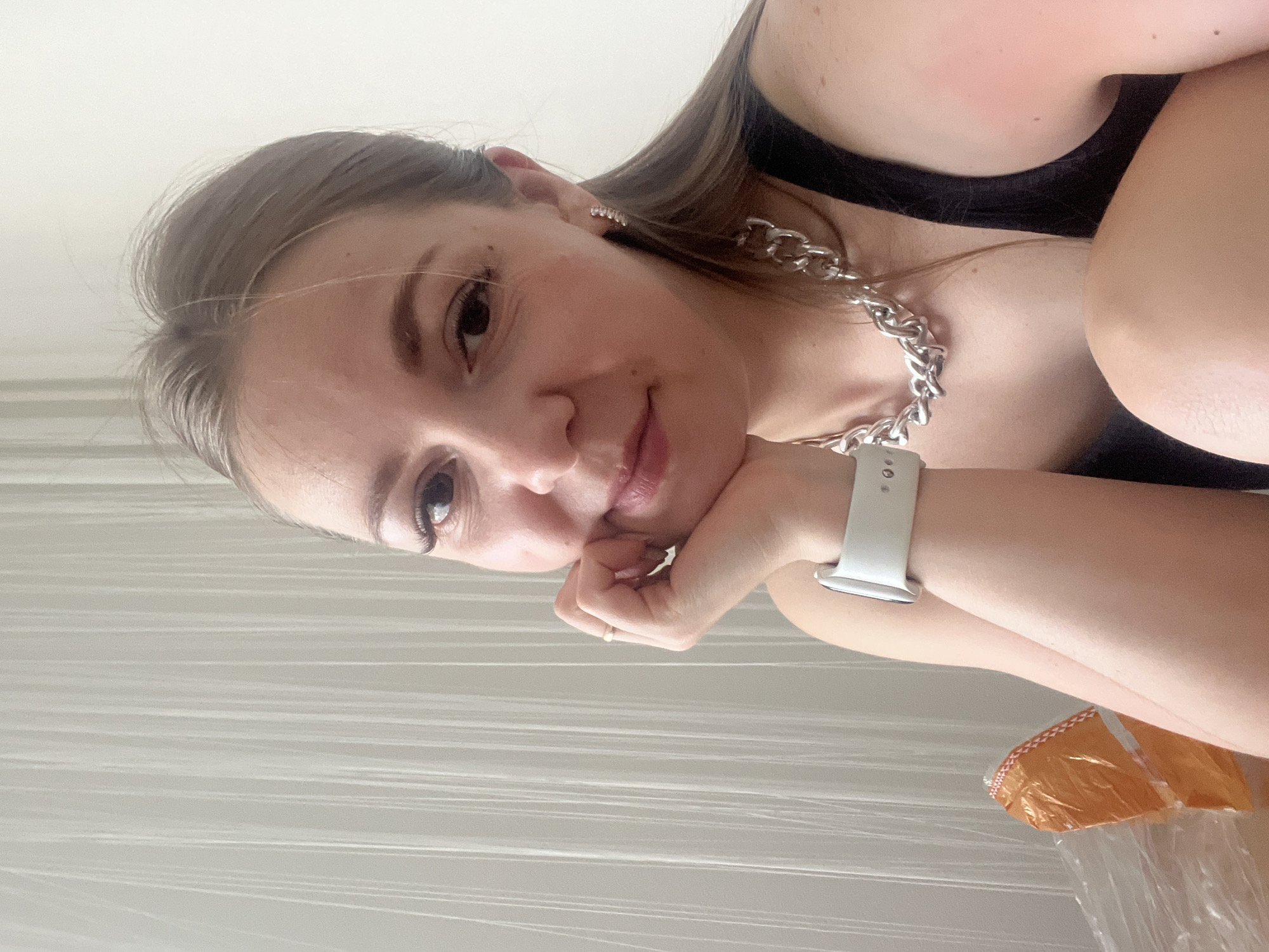 A young woman is pictured in a close-up, selfie-style portrait. She is wearing a black tank top, a thick, silver chain necklace, and a light-colored smartwatch on her left wrist. Her right hand rests against her chin, and she is wearing a thin gold ring on her right index finger. She has long, brown hair pulled back and is wearing small, sparkly earrings. She is looking at the camera with a slight smile. The background consists of a cream-colored wall and a sheer, white curtain with vertical pleats. Part of an orange package wrapped in plastic is visible in the lower left corner. There is no indication of the city of Chelyabinsk, Russia in the image.
