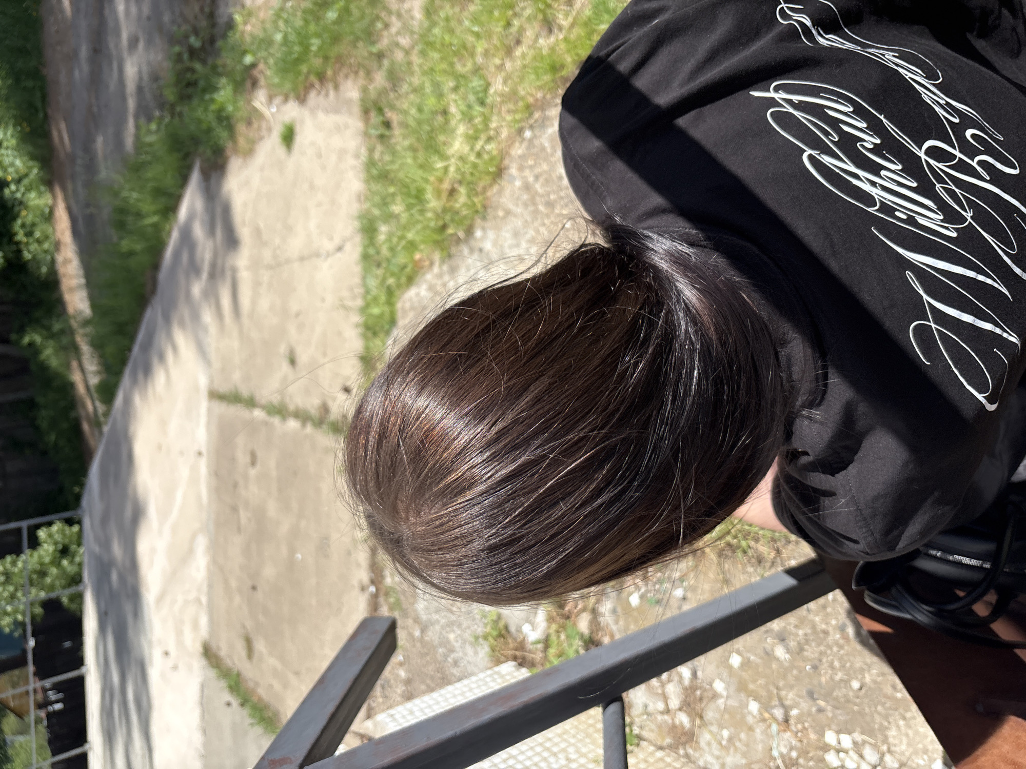 A high-angle close-up shot captures a person's head and shoulders. The person has dark brown hair with subtle highlights, appearing shiny. They are wearing a black hooded shirt with white script-like graphic designs on the back. The person is positioned near a gray metal railing with a concrete surface and grass visible in the background. The photograph appears to have been taken outdoors in daylight. The image does not contain any information to confirm the location as Chaikovsky, Russia.