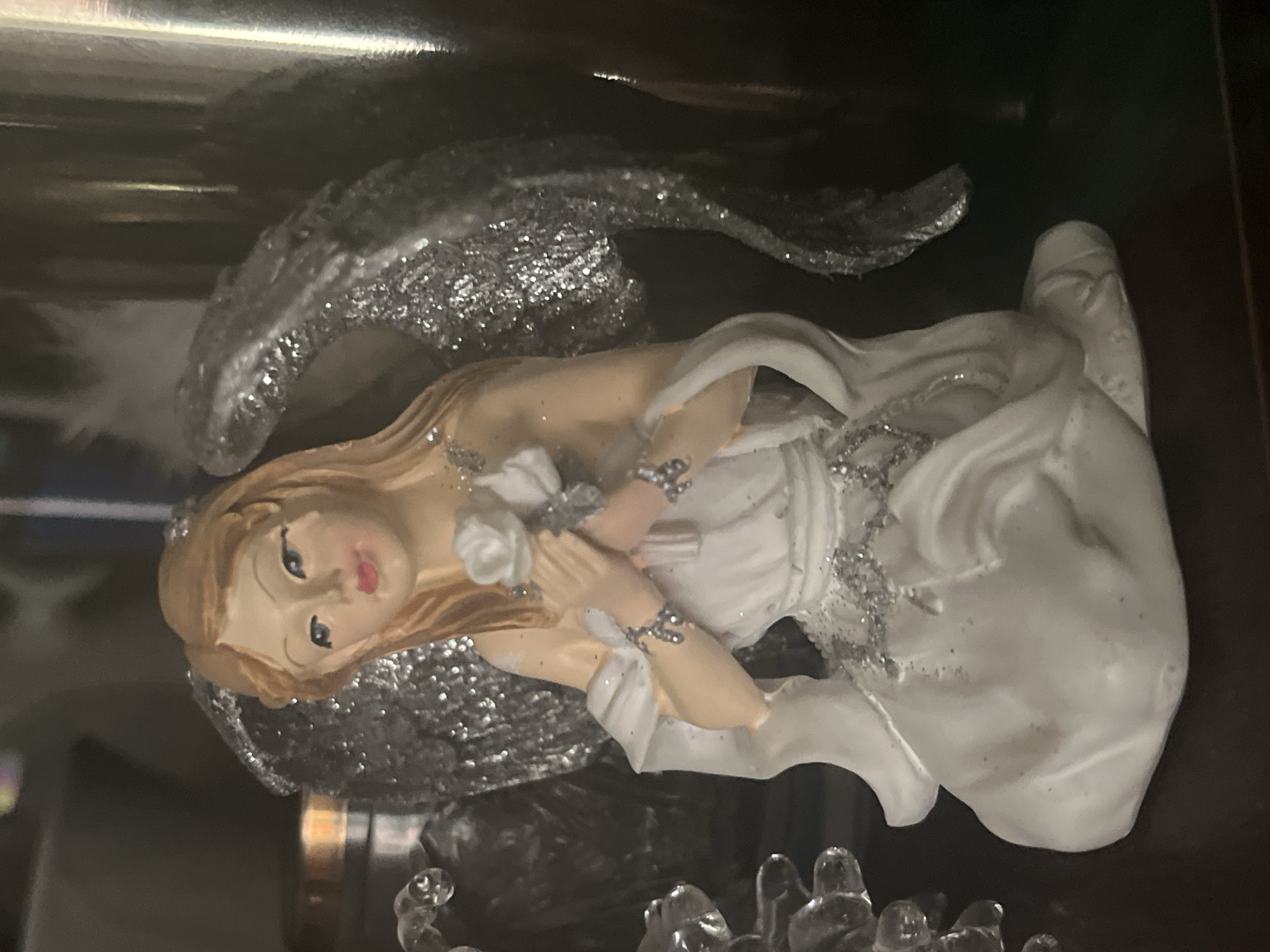 This is a close-up, eye-level, indoor photograph of a decorative angel figurine. The figurine is light-skinned with blonde hair, blue eyes, and pink lips. It is wearing a white, flowing dress adorned with silver glitter. The angel has large, shimmering silver wings. Its hands are clasped together, holding a white rose. The figurine is kneeling. To the left of the angel, there are some clear, glass-like decorative objects. The background is dark and out of focus, with some metallic and dark reflective surfaces visible. The lighting is soft, creating a warm glow on the figurine. The image does not contain any specific geographical markers that would indicate a location in Minsk, Belarus.