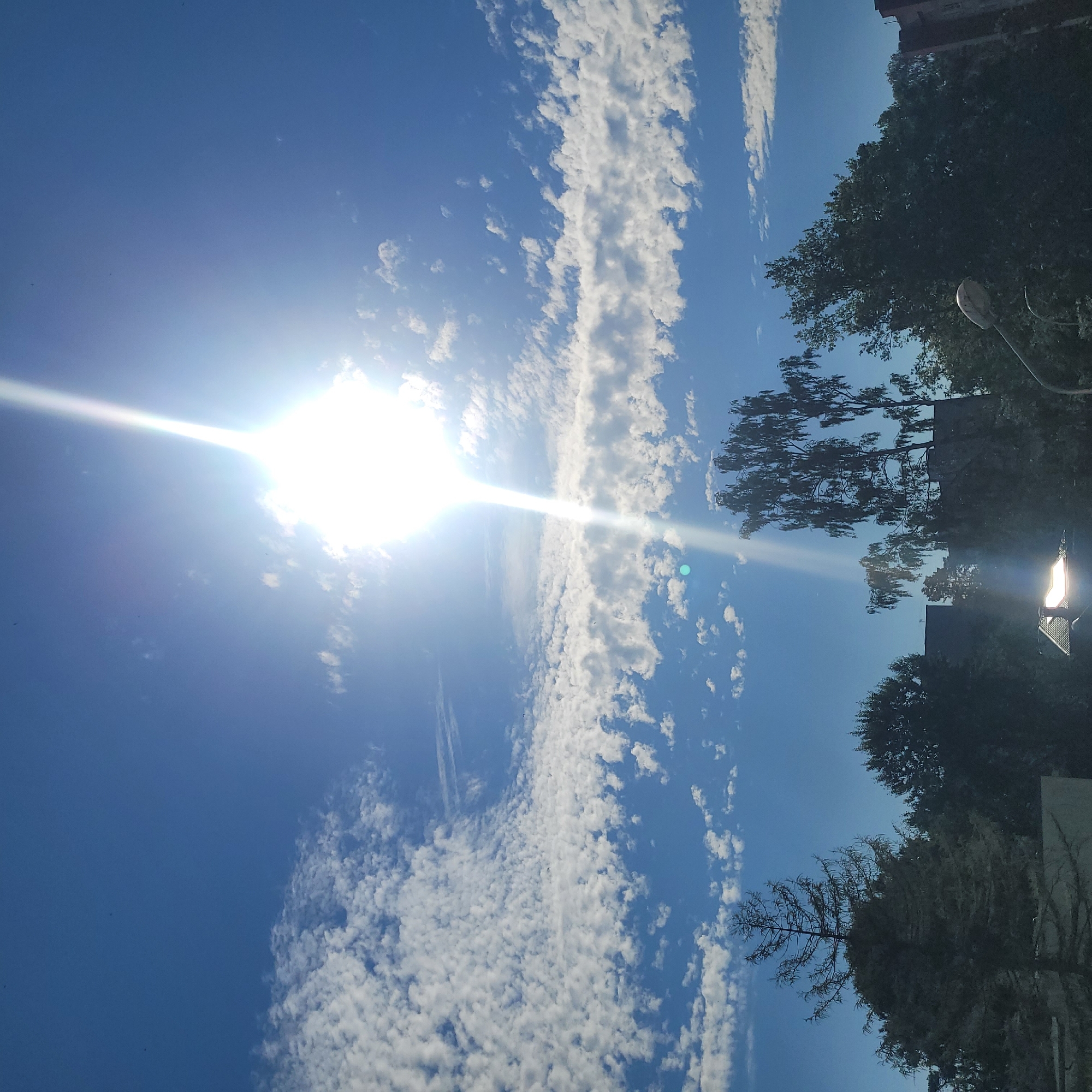 The image depicts a bright, sunny day in Jūrmala, Latvia, with the sun prominently featured in the upper center of the frame, casting a strong glare. The sky is a vibrant blue, scattered with fluffy white clouds that form distinct bands across the scene. In the foreground, a silhouetted line of trees and buildings runs along the bottom edge, with some buildings partially obscured by foliage. A streetlamp with a distinctive curved arm is visible in the lower right corner. The overall impression is one of a clear, warm day, likely during daylight hours.