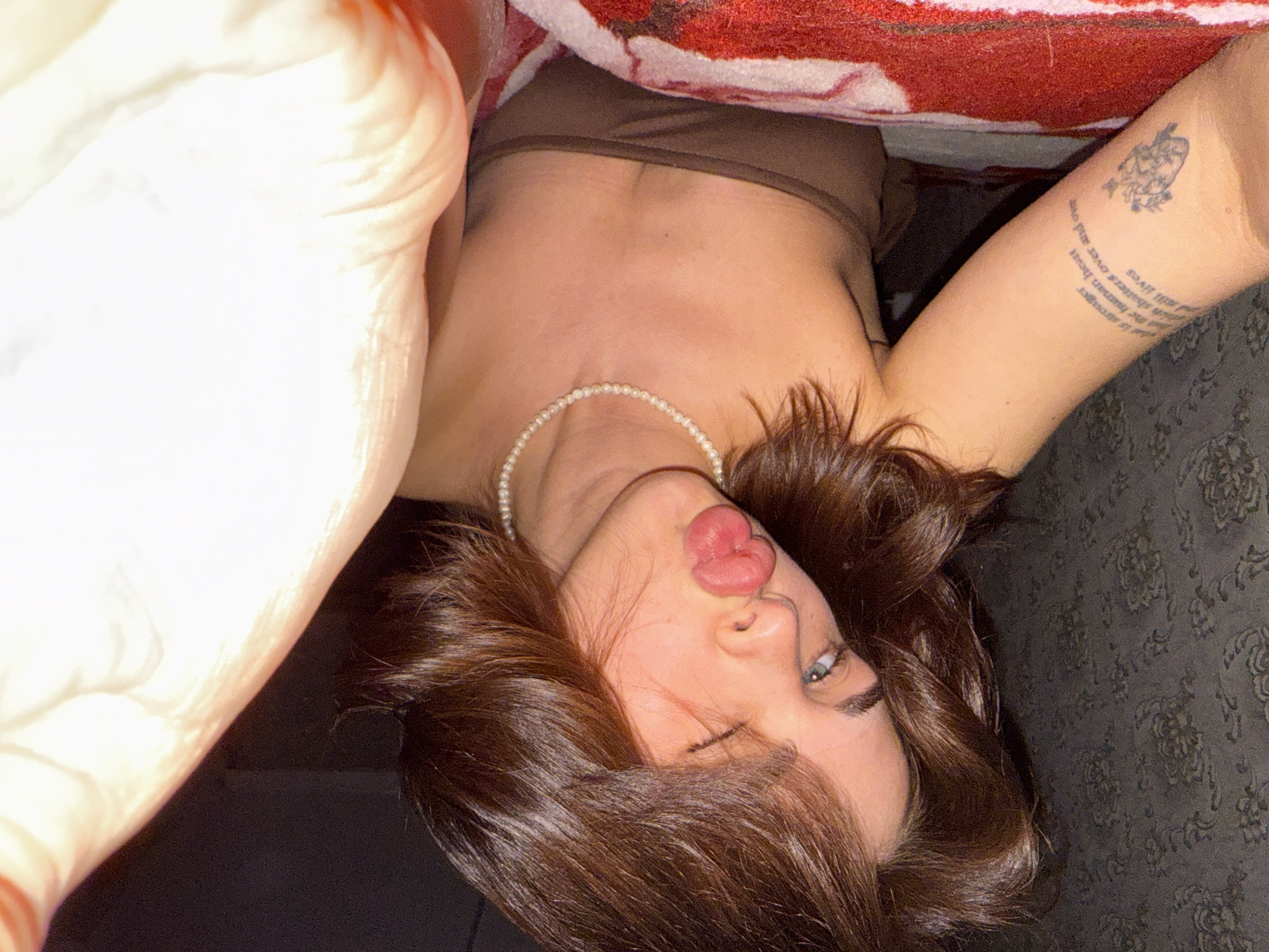 A woman lies on a dark floral patterned surface, possibly a bed or floor, indoors. She is partially under a red and white patterned blanket, wearing a brown top and a pearl necklace. Her brown hair is disheveled around her face, and she is making a kissing face towards the camera. The setting appears to be a private space. There is no information to identify the location as Sarapul, Russia.