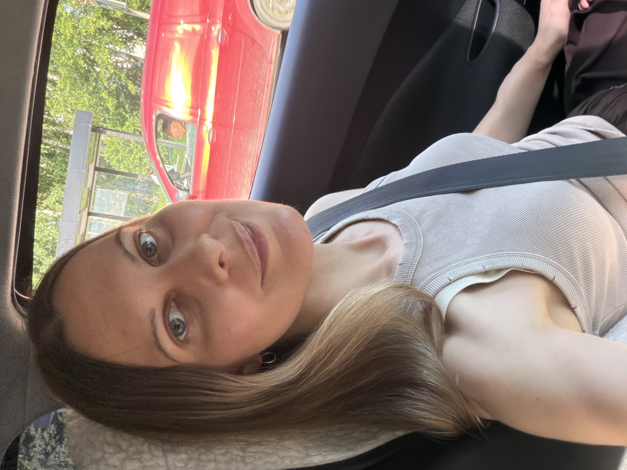 The media file shows a close-up, sideways selfie of a woman inside a car. She has blonde hair and is wearing a light-colored, ribbed top. A black seatbelt is visible across her chest. The setting appears to be daytime, with sunlight reflecting off a red car visible through the car window, which is to the left of the frame. Green trees and foliage are also visible outside the car, suggesting an outdoor setting. The woman's expression is neutral, and the overall mood of the image is casual. The orientation of the image is rotated.
