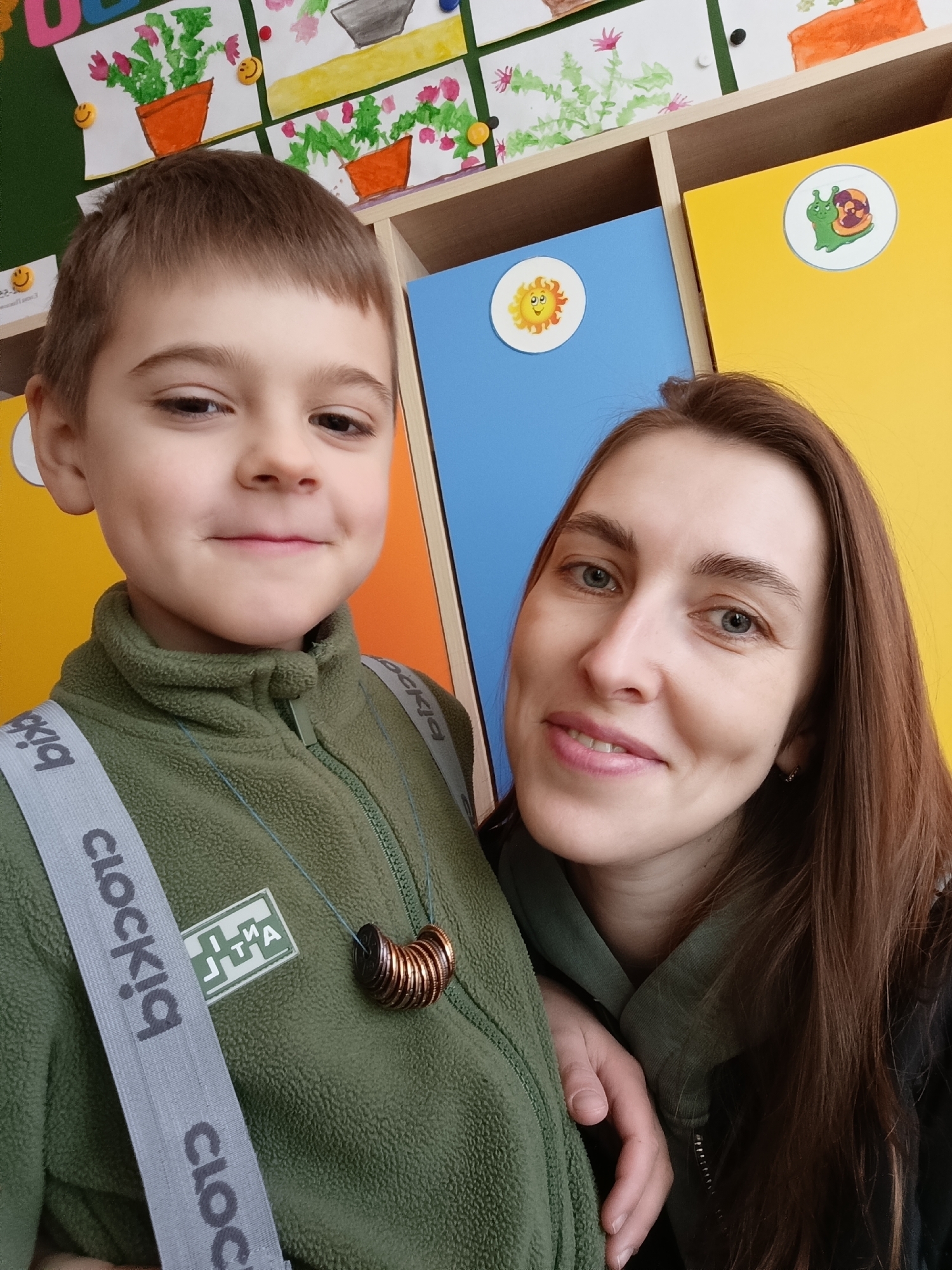 The image captures a selfie of a woman and a young boy, seemingly indoors, likely in a classroom setting, potentially in the city of Chaikovsky, Russia. The boy is positioned to the left of the frame, wearing an olive-green fleece jacket with grey suspenders bearing the word "clockiq". He also has a necklace made of stacked, dark brown circular objects. The woman is standing next to him, her auburn hair falling to the side. Behind them are yellow and blue lockers adorned with sticker decorations of a sun and a snail. Above the lockers is a shelf with potted plant drawings attached to the wall with colorful pushpins. The overall lighting appears natural, indicating daylight conditions.