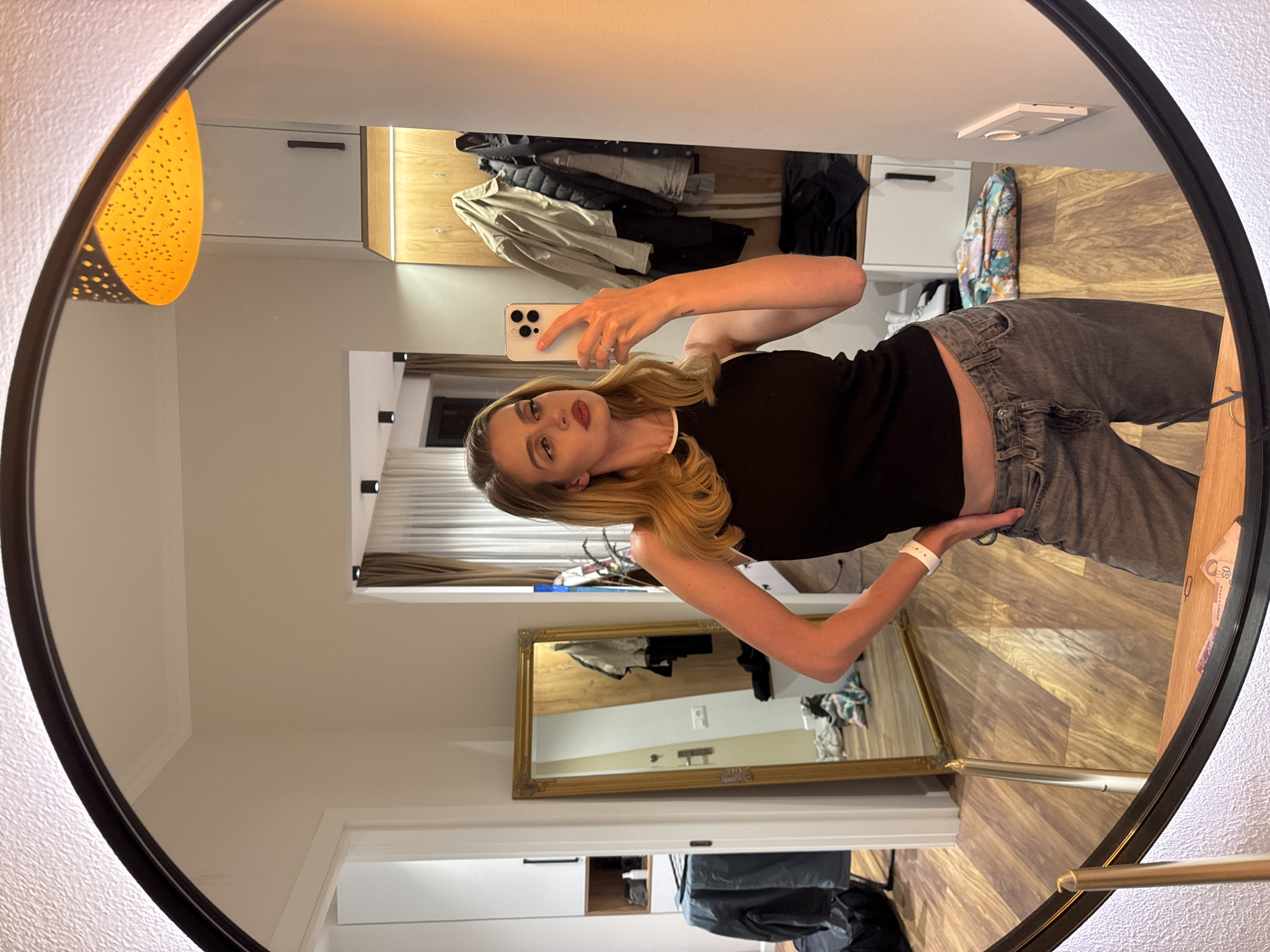 A young woman is captured in a mirror selfie, holding a smartphone up to her face with her right hand while her left hand rests on her hip. She has long, wavy blonde hair and is wearing a black sleeveless top with a white trim and grey jeans. A white wristband is visible on her left wrist, and a small ring is on her left ring finger. A faint tattoo, possibly reading "Artes," is visible on her inner left forearm. She has red lipstick.
The image is a reflection in a large, round, black-framed mirror. The setting appears to be an indoor room, possibly a dressing room or hotel room, with light-colored walls and warm-toned wooden flooring.
In the background, reflected in the mirror, there's a wardrobe or shelving unit with various clothes hanging, including a light-colored jacket and other dark garments. To the left, an orange-yellow light fixture with small perforations emits a warm glow. Another smaller, rectangular mirror with an ornate gold frame is visible on the wall behind her, reflecting more of the room's interior. On the floor to the right, some folded banknotes, showing "50," are visible on a wooden surface. A dark bin or bag is also partially visible on the floor in the reflection. The room has modern ceiling lights. The overall lighting is artificial and warm.