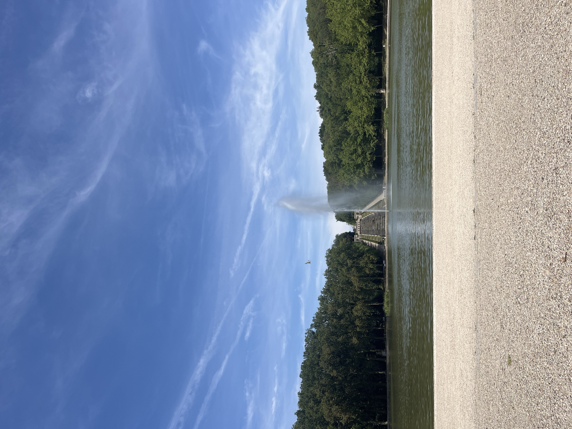 This is a horizontal, eye-level, medium shot of a large body of water in the Parc de Sceaux, located in Antony, France. The sky is bright blue with thin, wispy white clouds. In the center of the image, a tall fountain shoots water upwards from the lake. Beyond the lake, a dense line of dark green trees forms a backdrop, with a set of ornate stone stairs visible on the right side of the frame, leading up to the trees. In the foreground, a path made of light-colored gravel and small stones stretches across the bottom of the image. A single bird is in flight in the upper portion of the image, over the lake.
