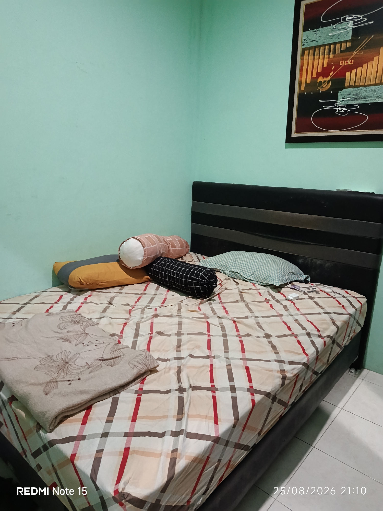 A bedroom scene in Keboananom, Indonesia features a bed with a patterned comforter and several pillows. The bed has a dark headboard. A framed piece of abstract art hangs on the light green wall above the headboard. The floor is tiled. A timestamp in the lower right corner indicates "25/08/2026 21:10". A watermark in the lower left corner reads "REDMI Note 15".