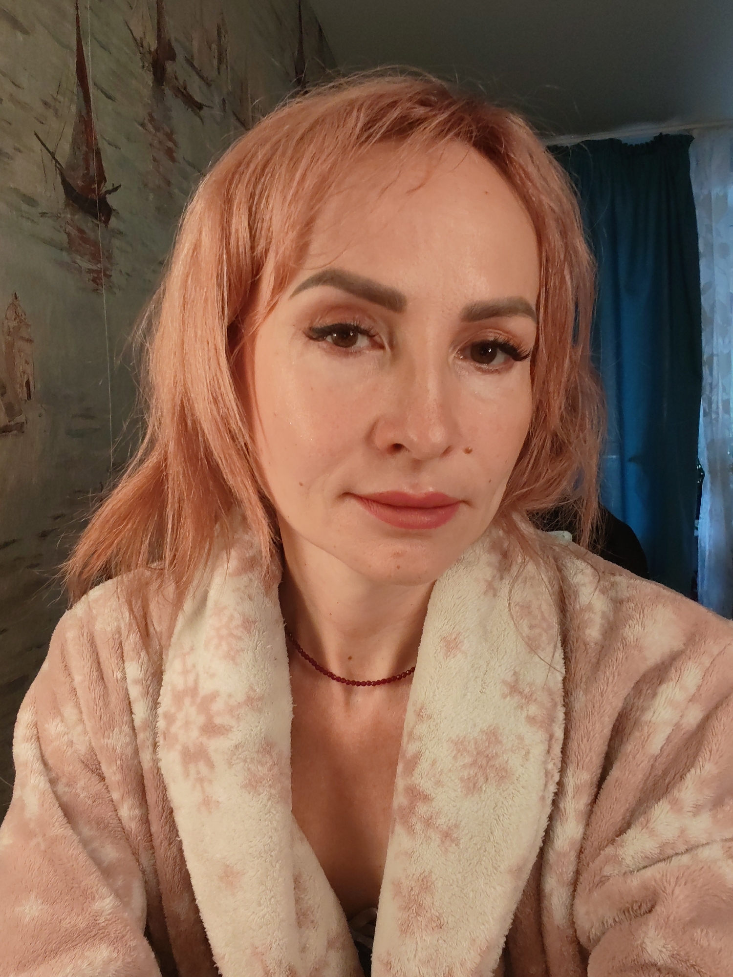 An indoor, eye-level, medium shot captures a fair-skinned woman with salmon-pink hair and light brown eyes. She is wearing a plush pink bathrobe with a snowflake pattern and a thin red beaded necklace. Her makeup includes subtle eyeshadow, eyeliner, mascara, and pink lipstick. The background consists of a patterned wallpaper with ships and water, and teal curtains. The lighting appears to be natural, coming from a nearby window. There is no indication of the city of Chaikovsky, Russia, in the image.