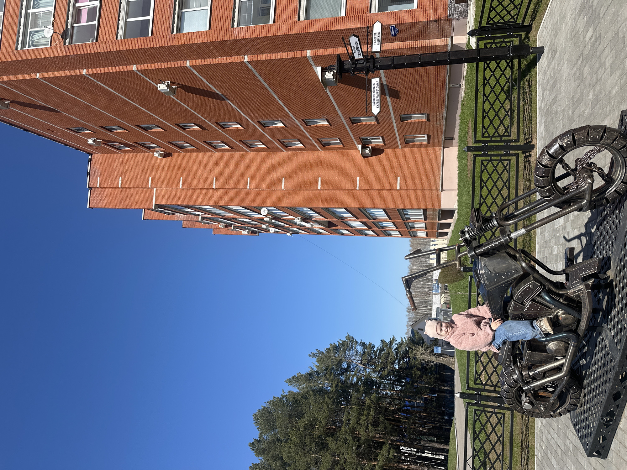 A young child wearing a pink jacket and blue jeans is sitting on a black metal sculpture of a motorcycle. Behind the motorcycle, there is a green fence and tall pine trees under a clear blue sky. Further back, a brick building with numerous windows and satellite dishes is visible. A street sign post stands near the building. This scene is in Chaikovsky, Russia.